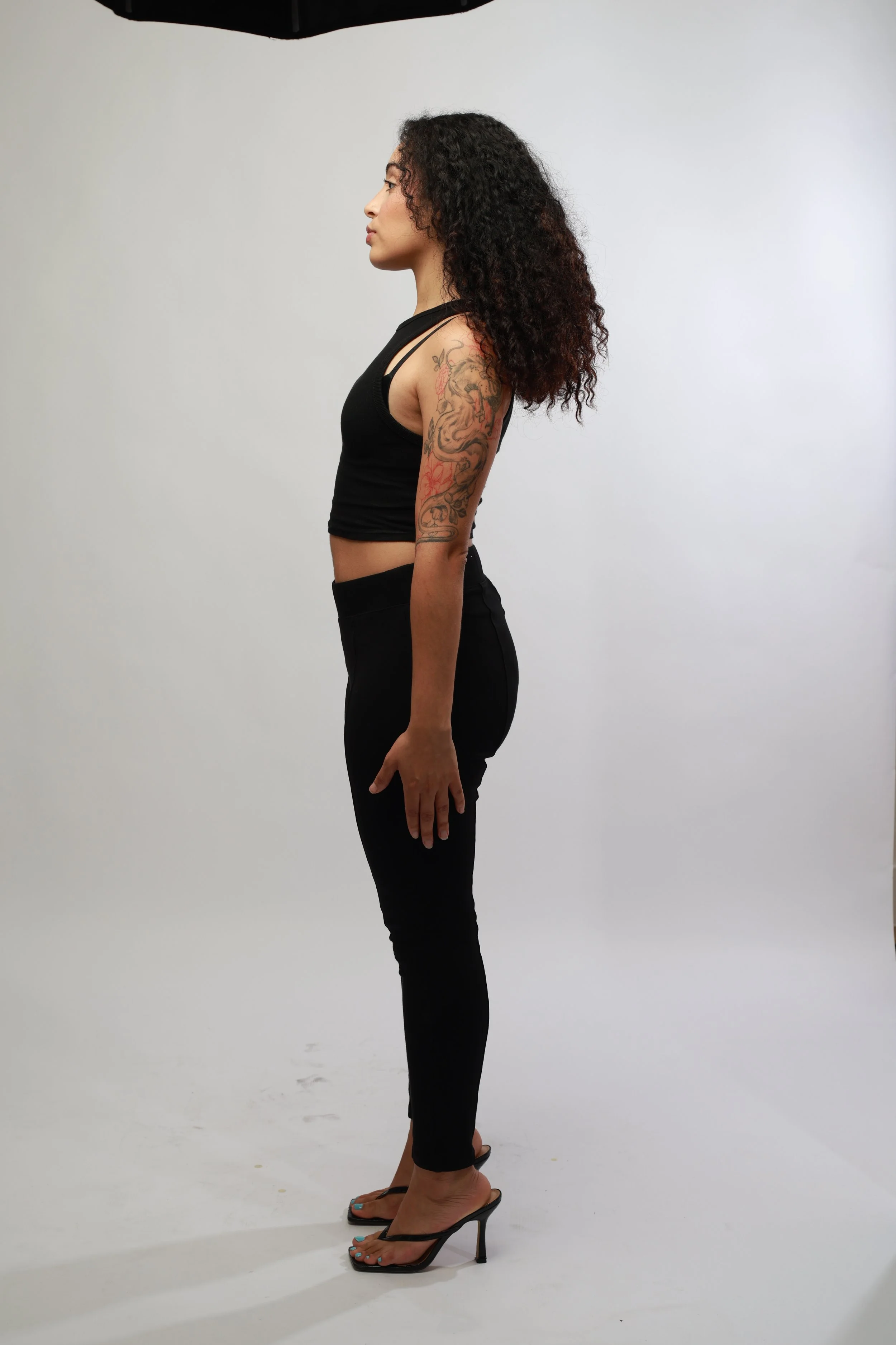 Side profile of a woman with curly dark hair, wearing a black sleeveless crop top, black pants, and high-heeled sandals, standing against a plain white background.