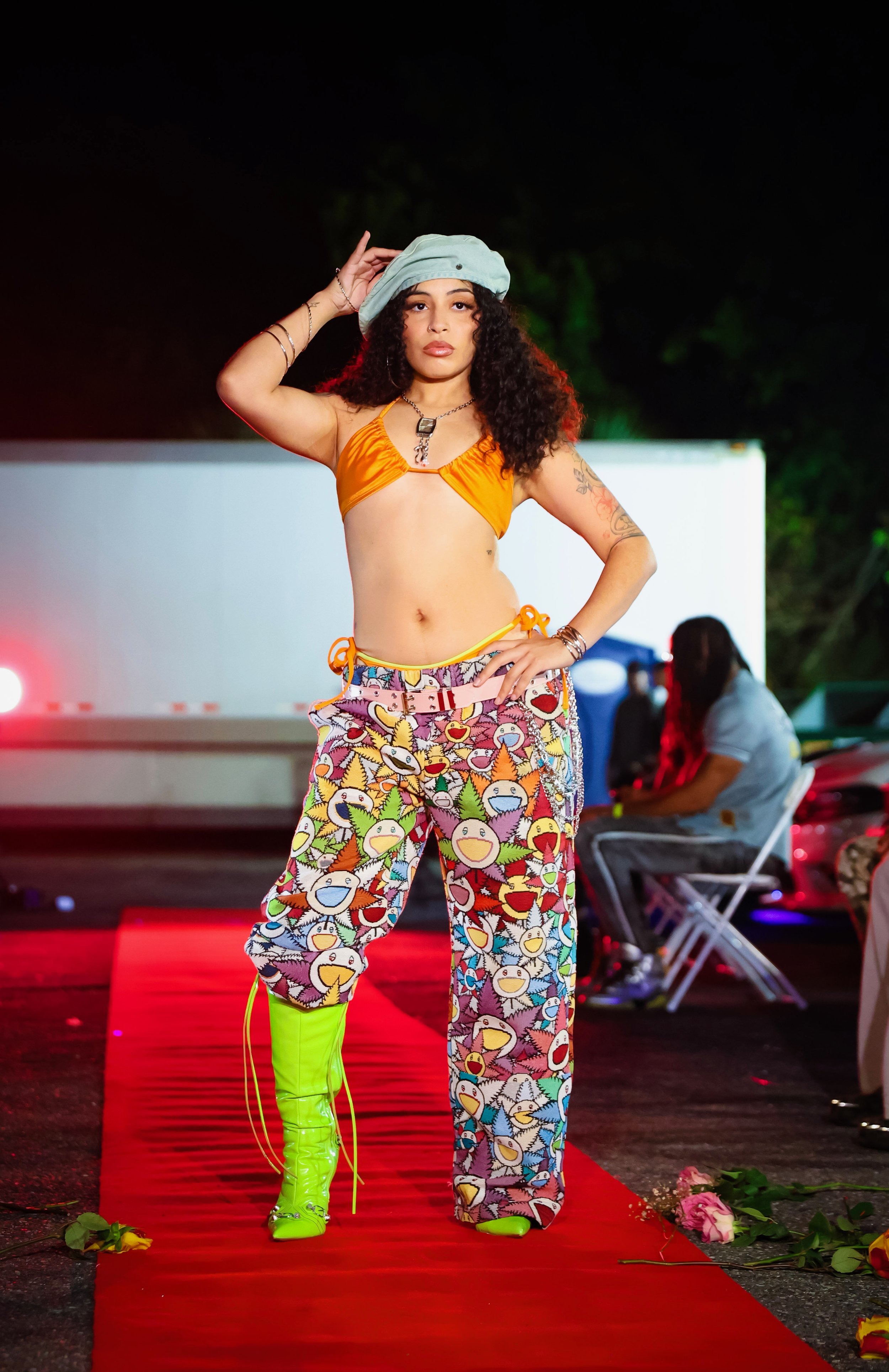 A woman standing on a red carpet at night, wearing bright green thigh-high boots, colorful pants with cartoon faces, an orange bikini top, a light blue hat, and accessories, with a flower and rose petals on the ground nearby, and other people in the background.
