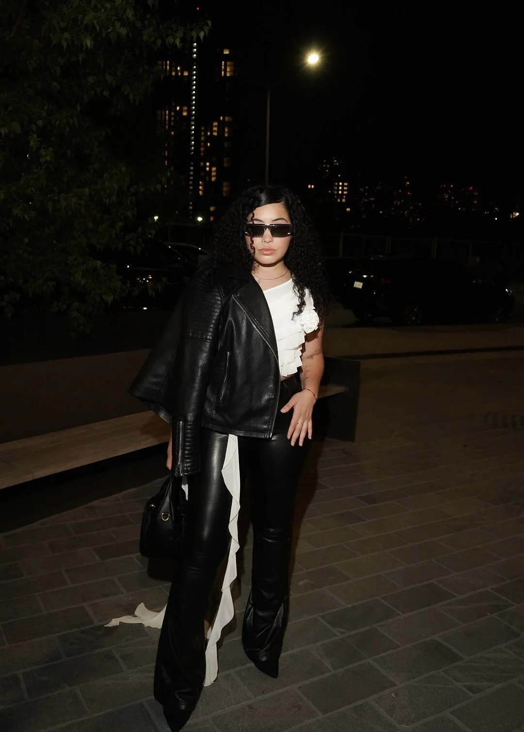 A woman with curly black hair wearing sunglasses, a black leather jacket draped over her shoulders, a white top with ruffles, black leather pants with a white stripe, standing on a city sidewalk at night.