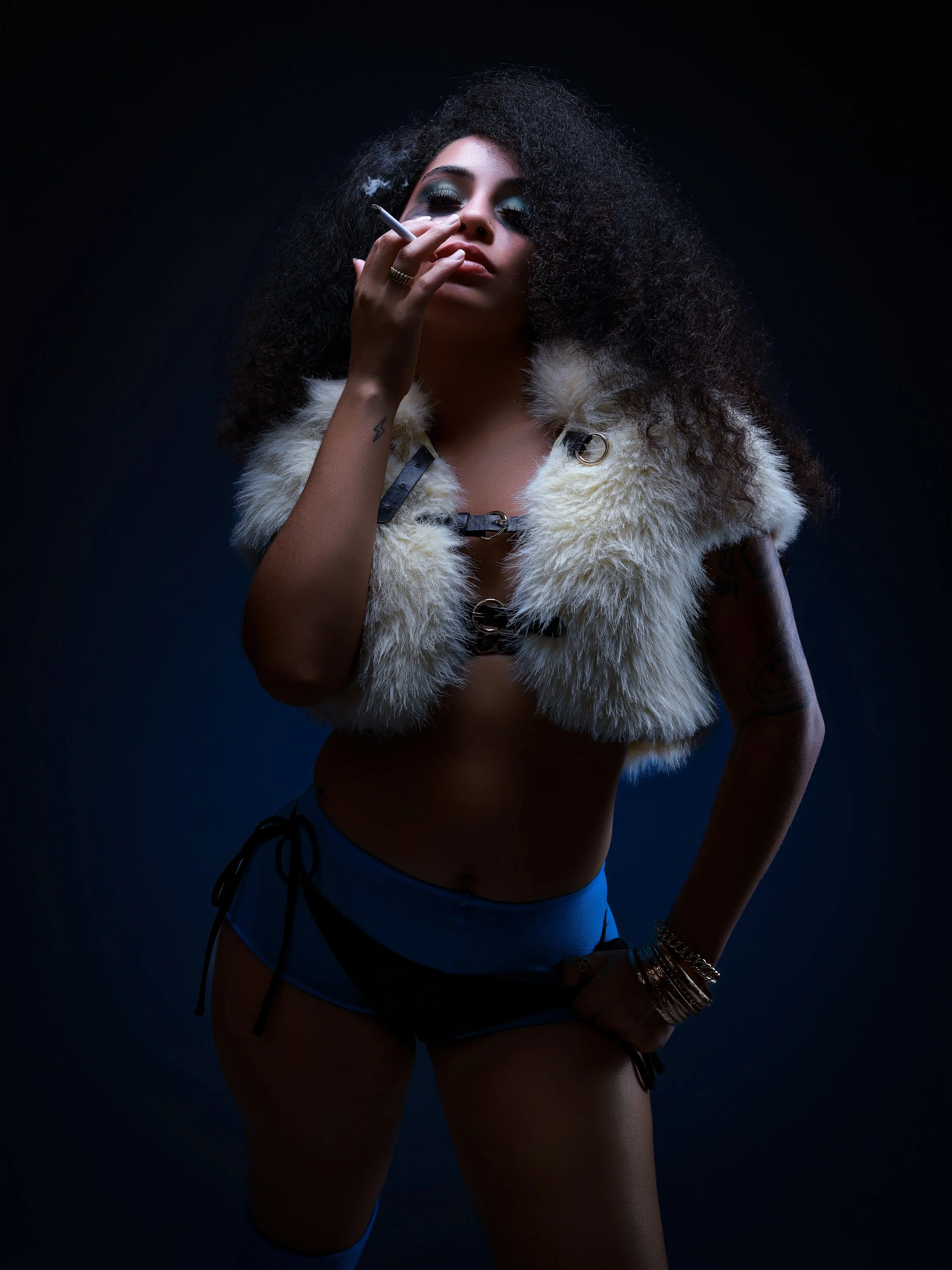 A woman with curly hair and dark makeup wearing minimal clothing, a fur vest, and multiple bracelets, holding a cigarette against a dark background.