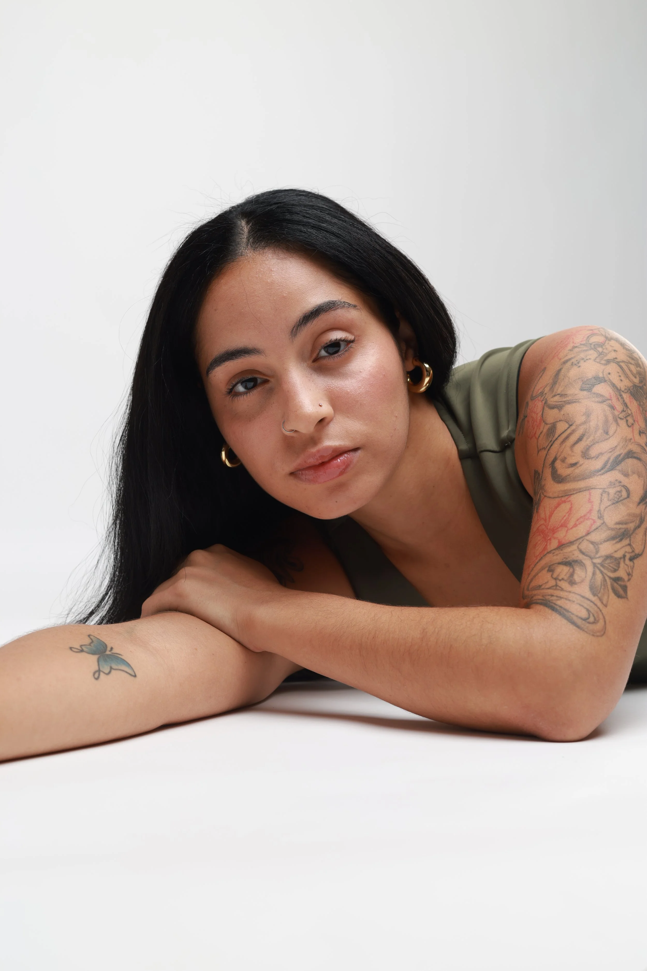 A young woman with black hair, wearing hoop earrings and tattoos on her arms, is lying on her side on a white surface against a light background.