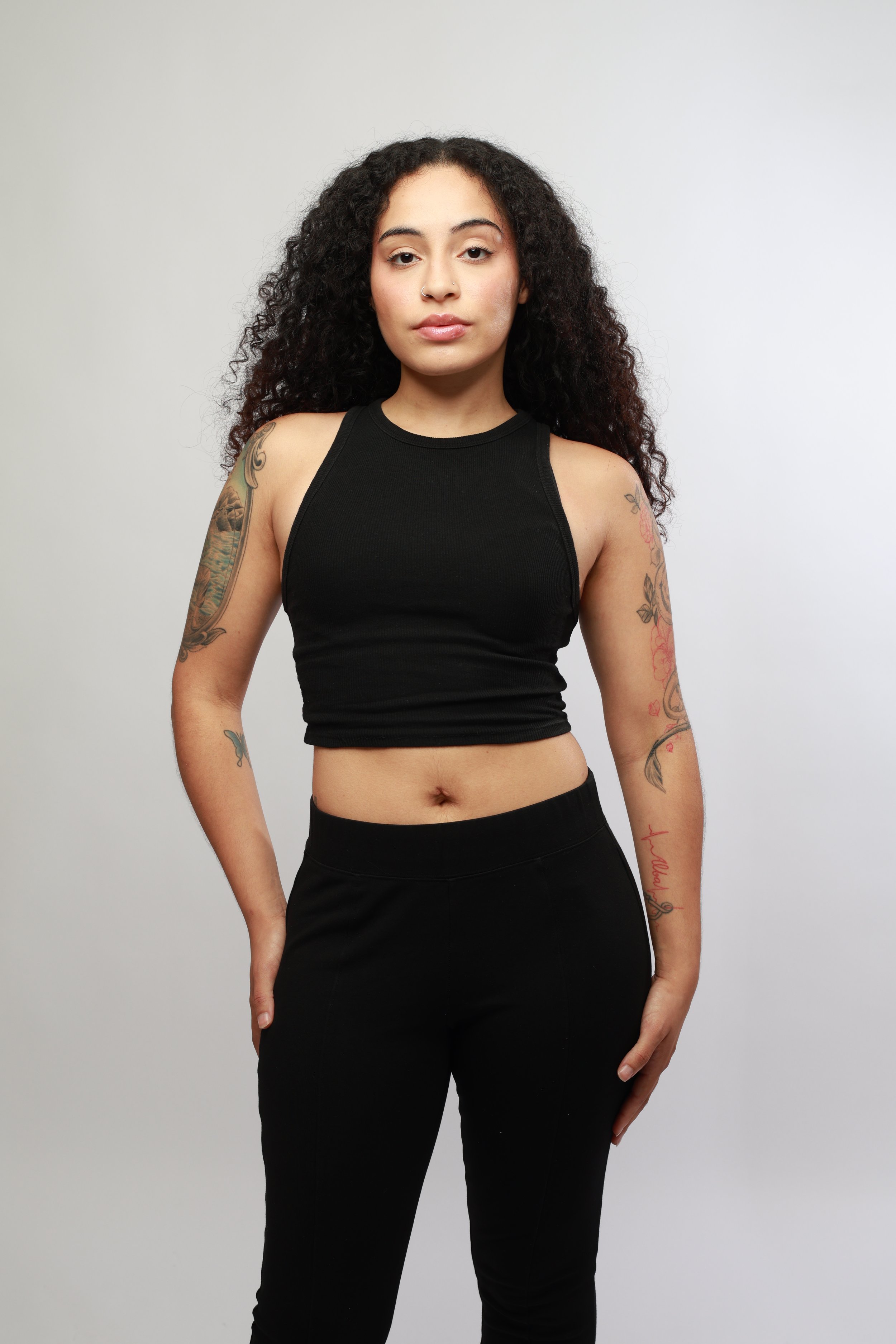 A young woman with curly dark hair and tattoos on her arms, wearing a black sleeveless crop top and black pants, standing against a plain light gray background.