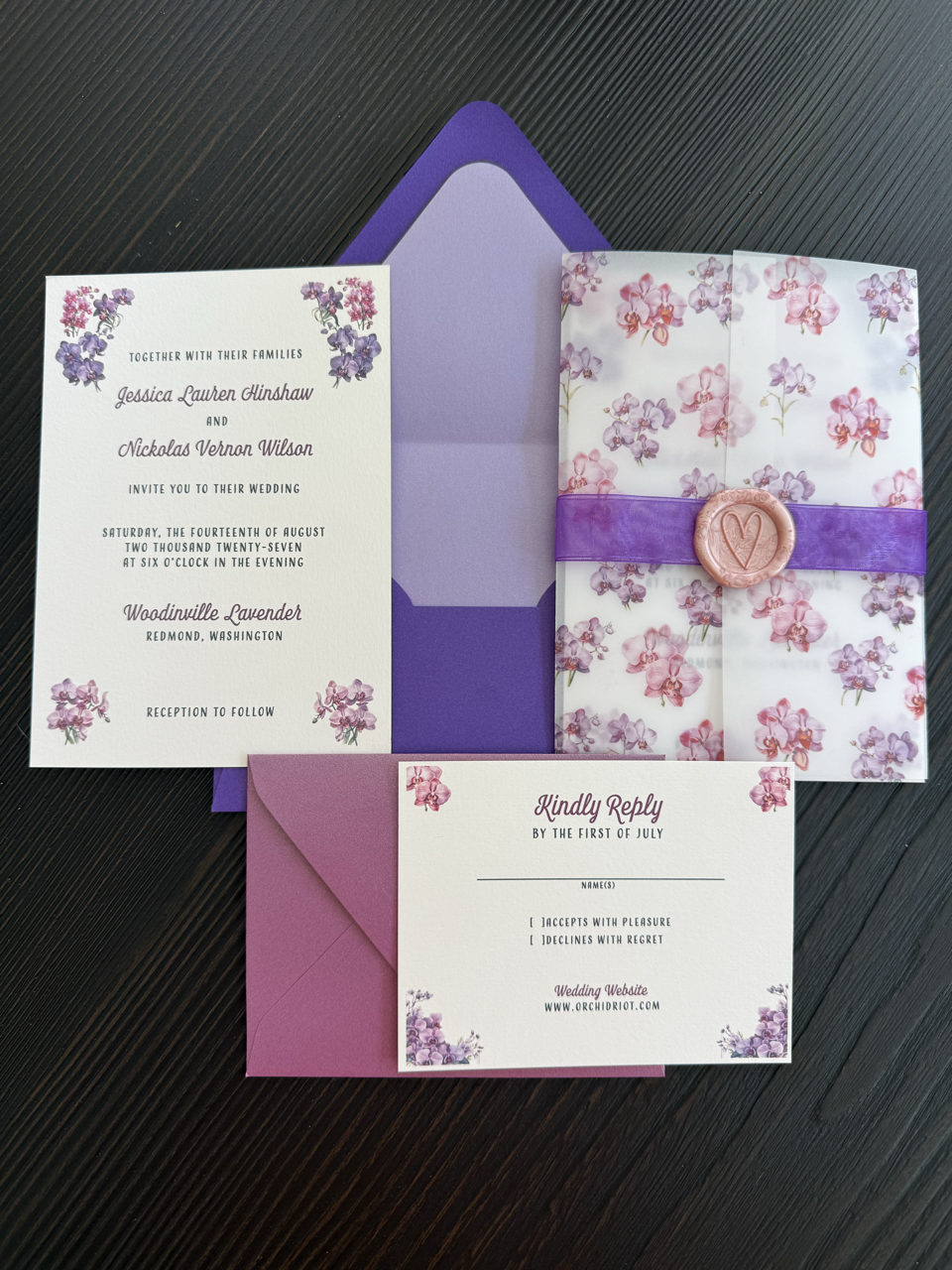 A wedding invitation set on a dark wooden surface. The set includes a white card with purple and pink floral designs, purple envelope, a pink reply card with floral accents, and a wrapped gift with floral paper and a purple ribbon with a wax seal.