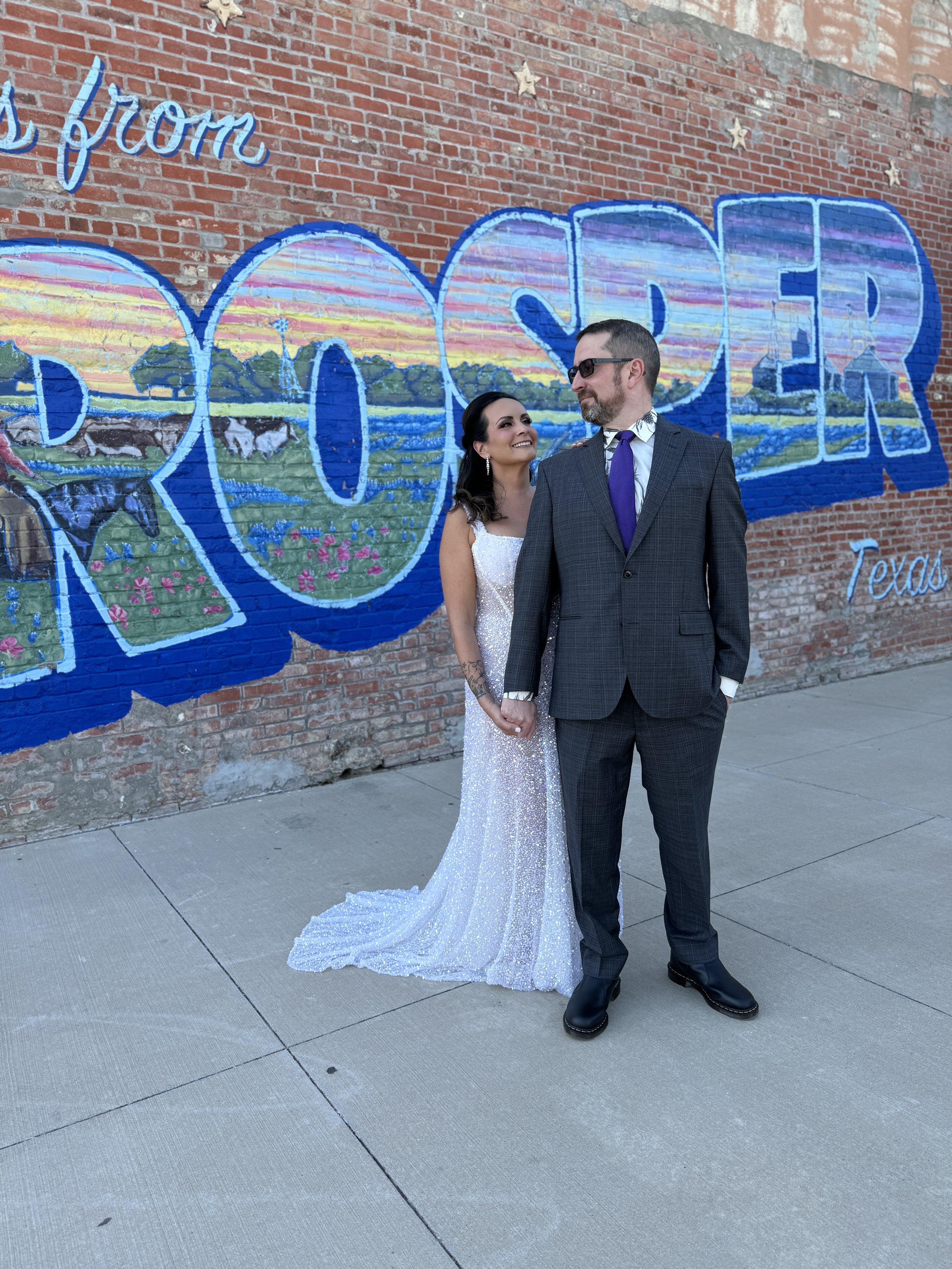 A couple in wedding attire holding hands and smiling while standing in front of a city mural that says "DALLAS" on a brick wall.