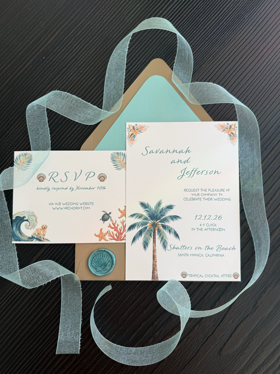 Beach themed wedding invitations with palm trees, seashells, starfish, fish, waves, and a dog illustration, along with RSVP card and blue seal, on a dark wooden surface with a sheer ribbon.