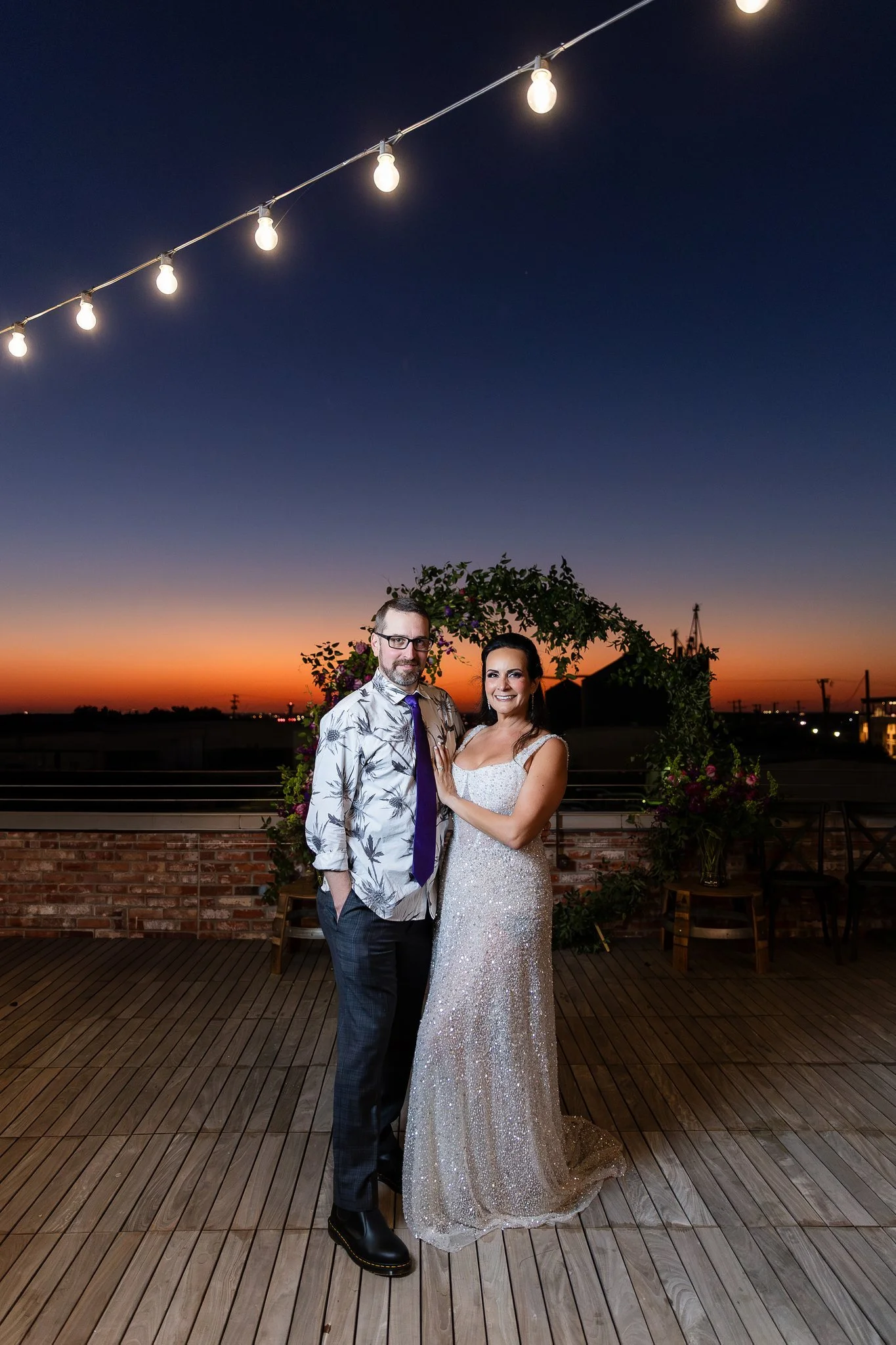 A couple stands close together on a rooftop deck at sunset, with string lights overhead and a floral arch behind them. The man is wearing glasses, a floral shirt, and dark pants, while the woman is in a sparkling wedding gown and smiling at the camera.