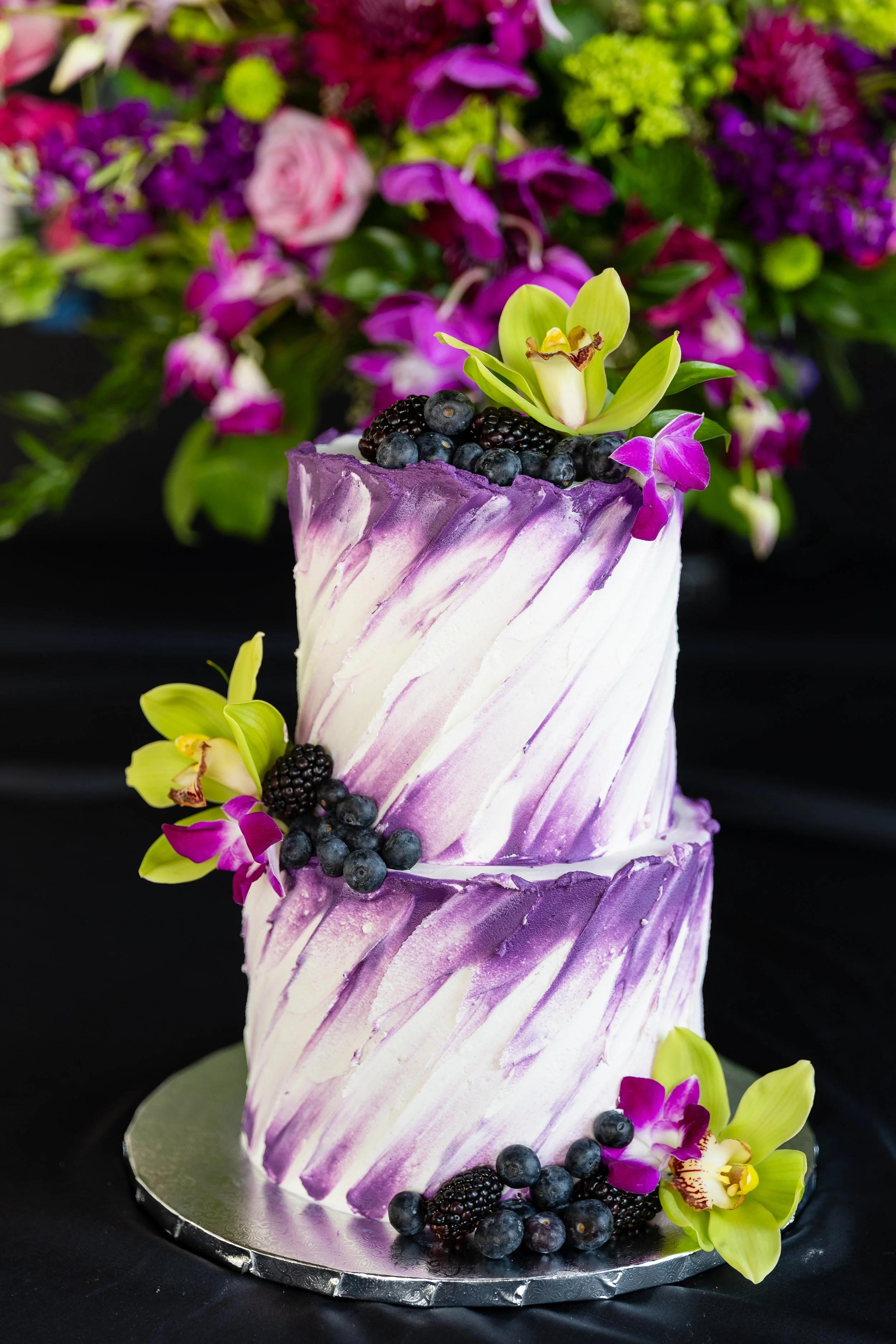 A tall, elegant cake with a purple and white marbled icing design, topped with blackberries, blueberries, and yellow and purple flowers, surrounded by matching flowers on a black background.