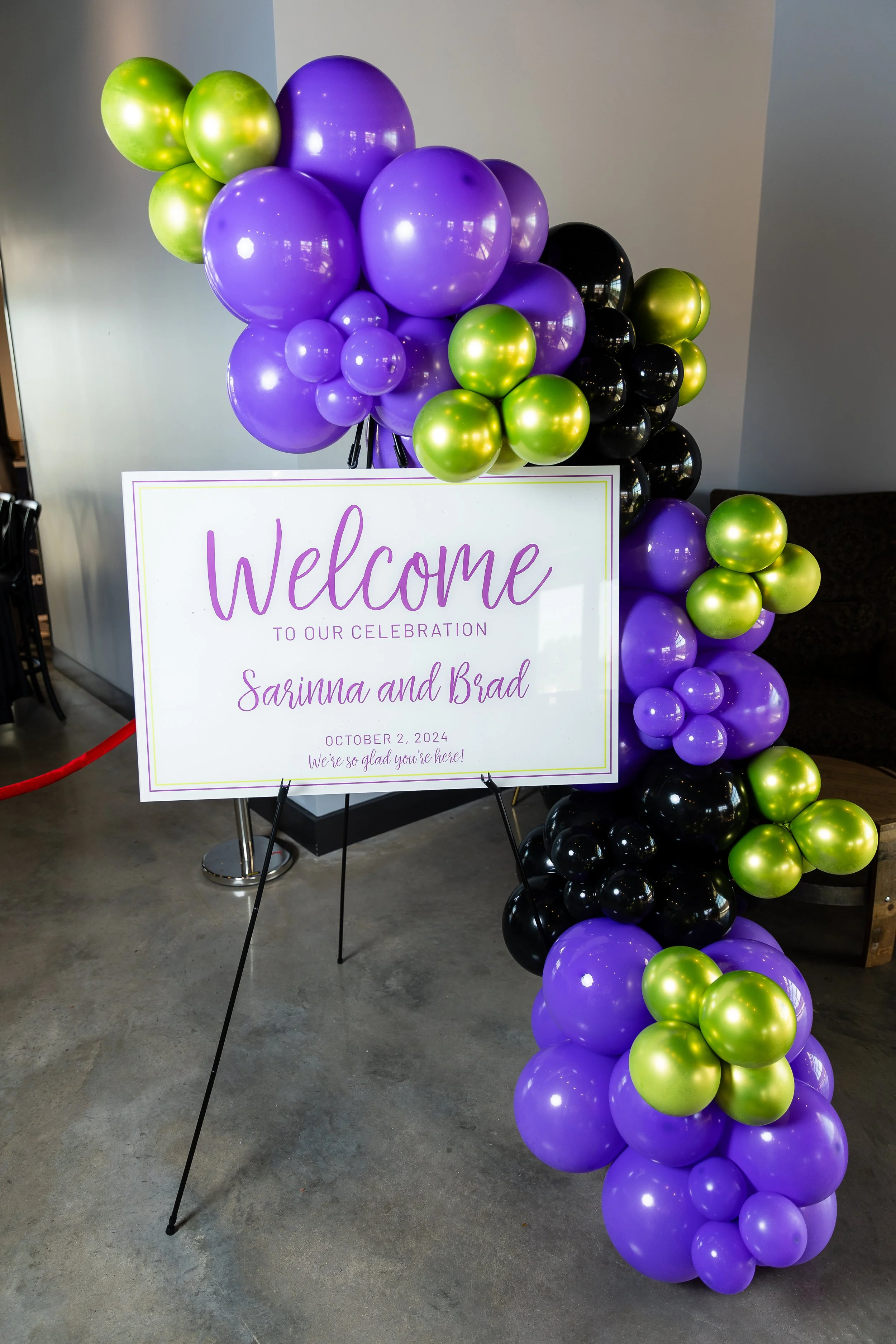 A welcome sign for a celebration featuring purple, black, and green balloons arranged in a decorative arch and cluster.