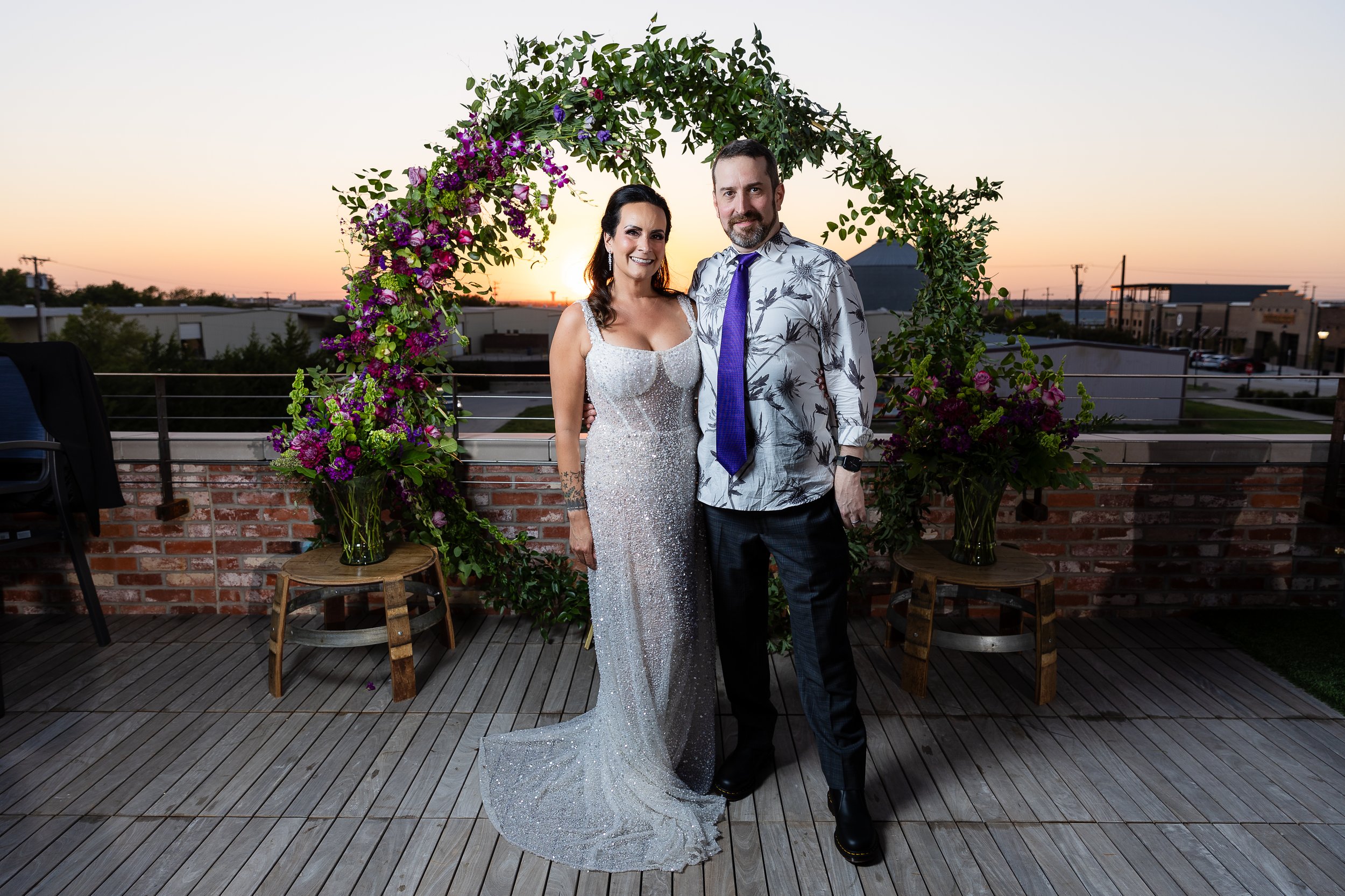 A smiling couple stands under a decorated floral arch during sunset on a rooftop terrace. The woman wears a white, form-fitting, sequined dress with spaghetti straps, and the man wears a patterned shirt with a purple tie and dark pants. There are small wooden tables with vases of flowers and chairs on the terrace, and a cityscape with buildings is visible in the background.