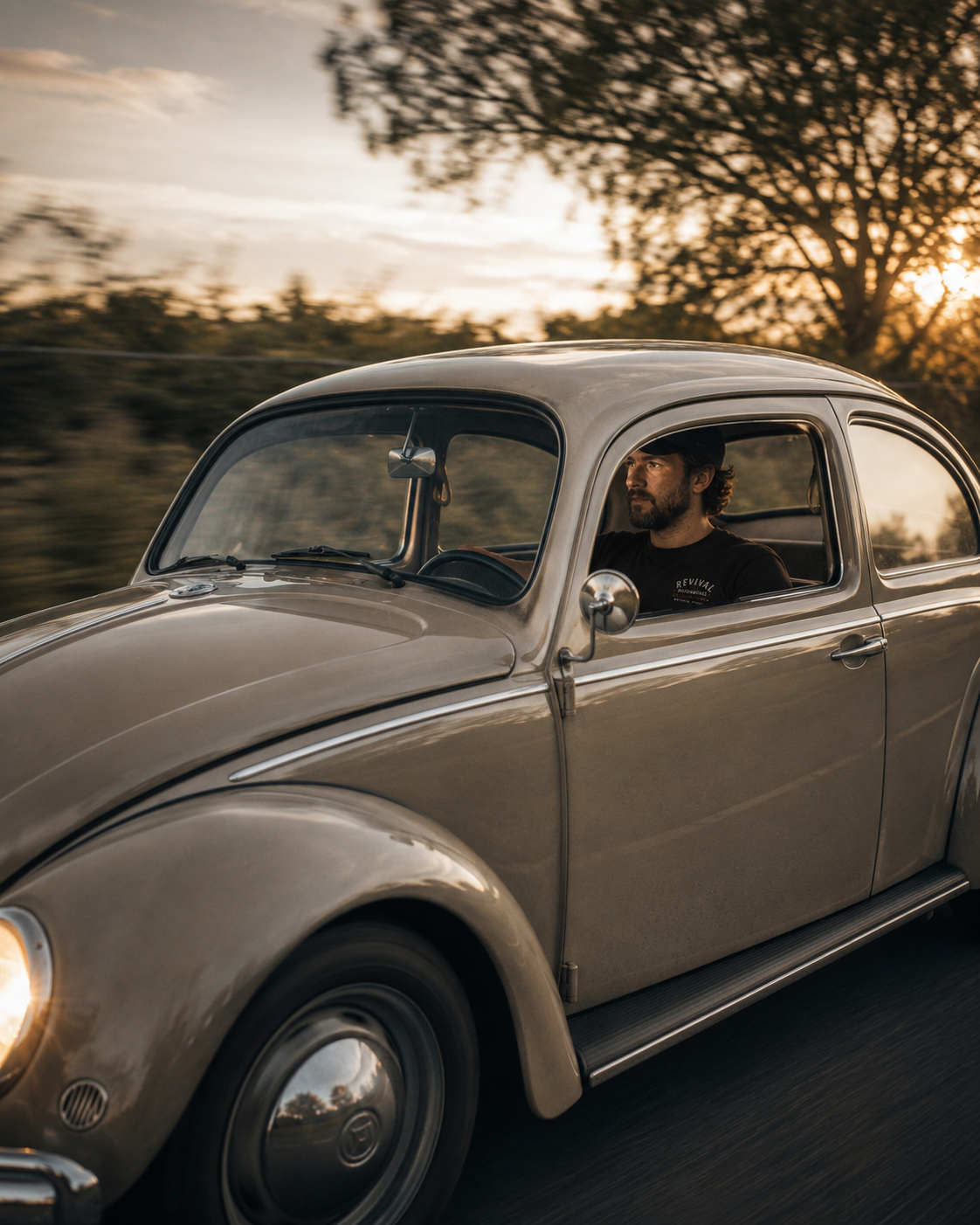 A man driving a vintage beige car during sunset with trees in the background.