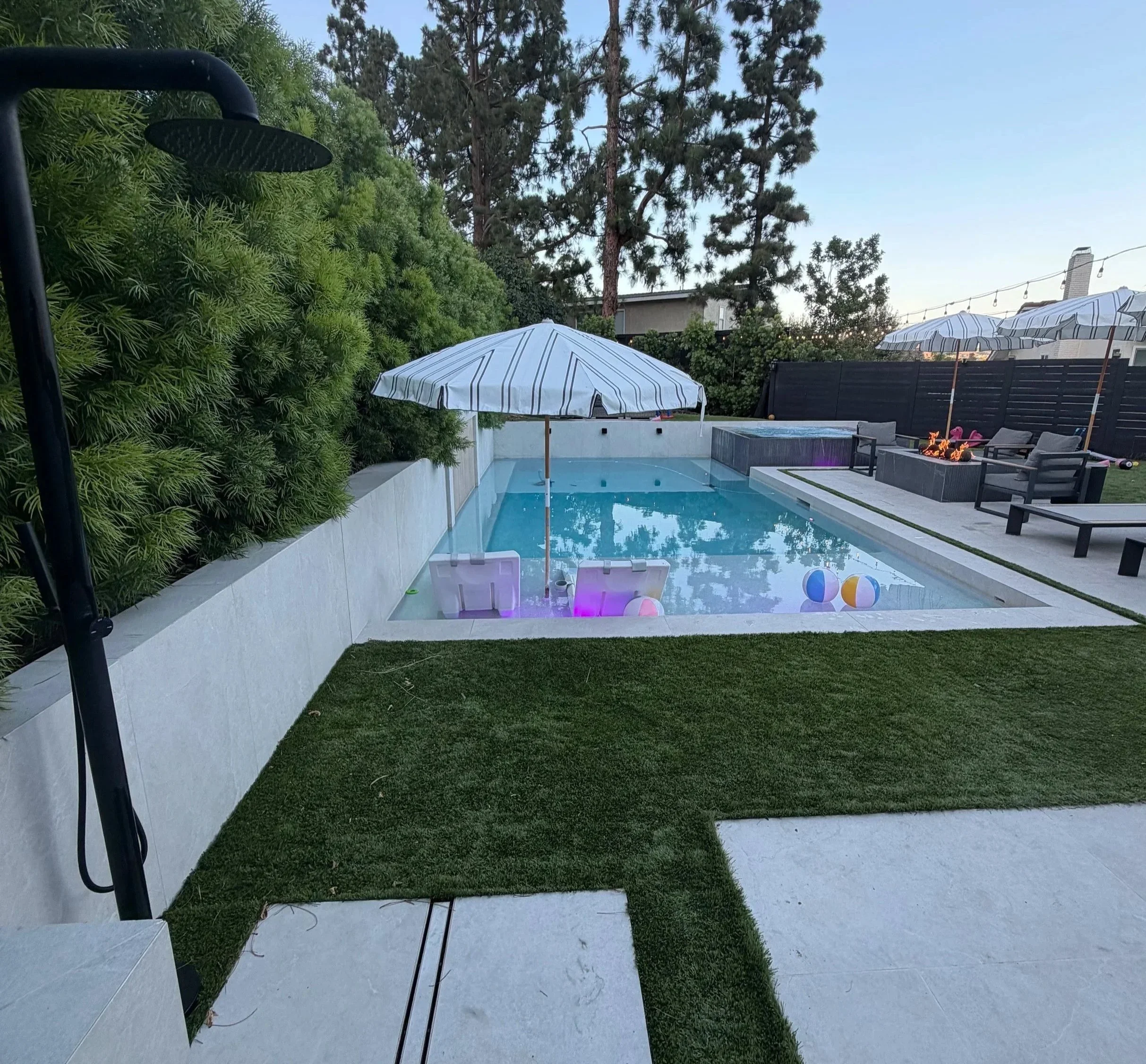 Backyard with a swimming pool, two patio umbrellas, outdoor seating area with firepit, string lights, surrounded by greenery and tall trees, during early evening. Services Offered: Bathroom remodel kitchen remodel room addition, whole house remodel