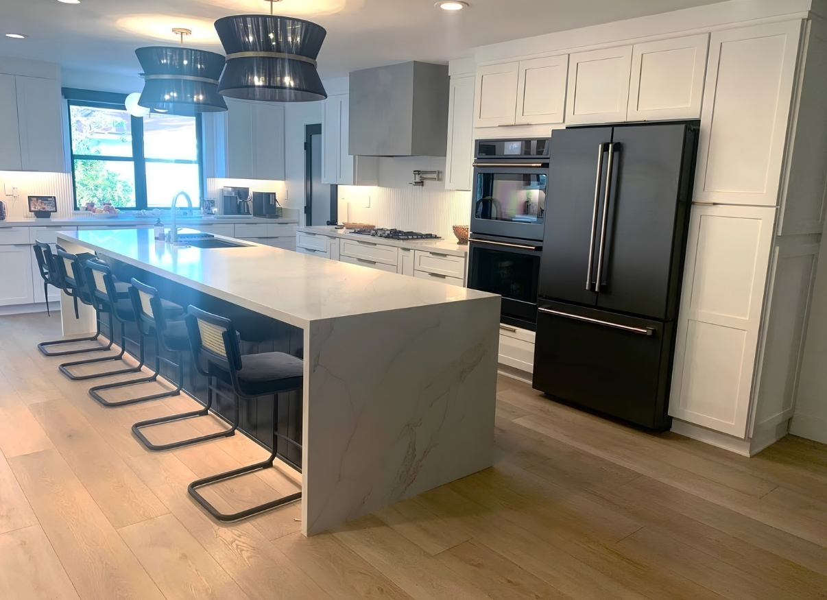 Modern kitchen with white wood and cabinetry, black modern appliances, a large quartz island with seating for five, and light wood floors and under counter lighting.  Masone Construction Inc. serves areas including Yorba Linda, Villa Park, Anaheim