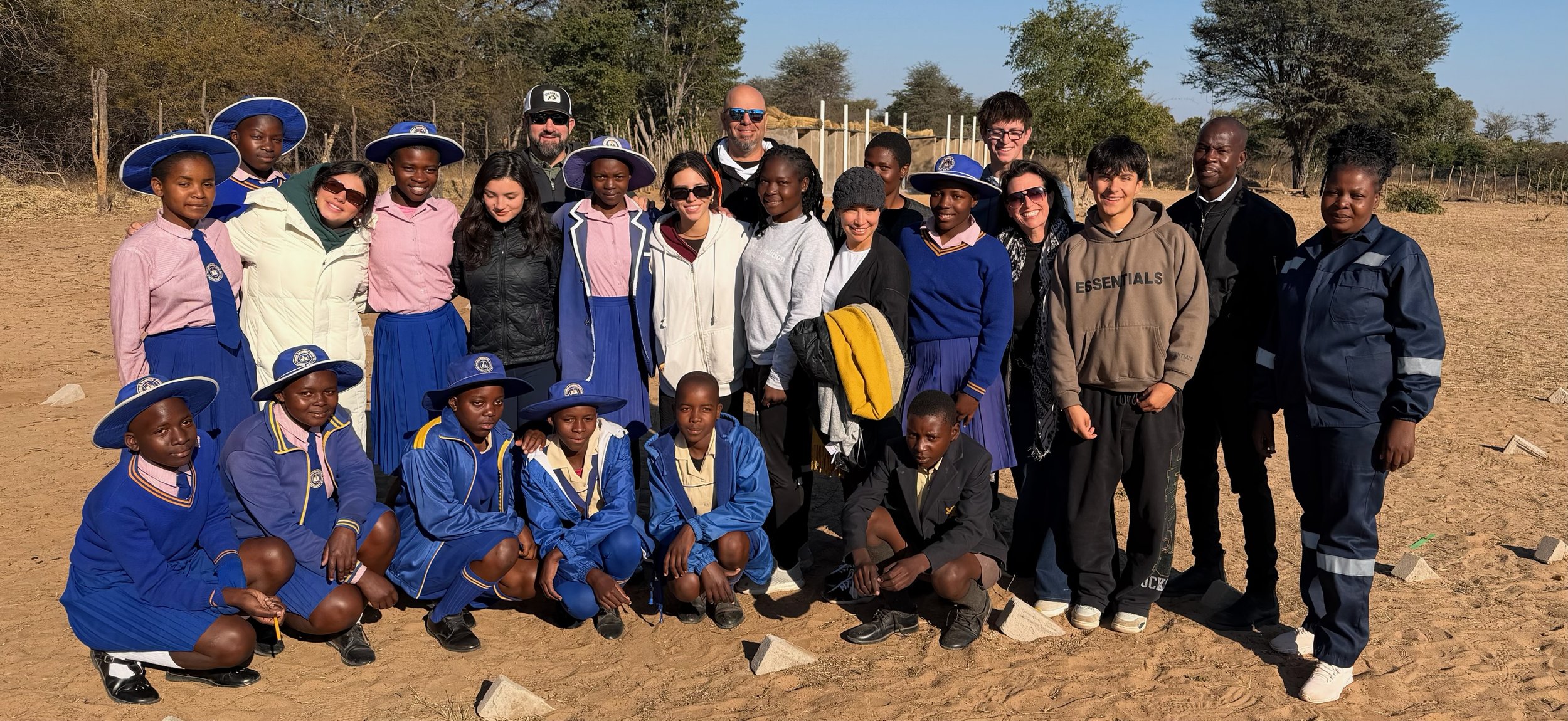 A group of children and adults posing outdoors on a sunny day, some children in school uniforms and others in casual clothing, with a dirt ground, trees, and a wooden fence in the background.