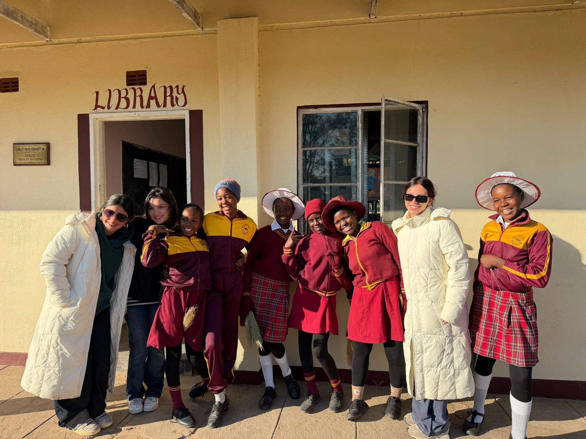 Group of children and two women standing outside in front of a building with a sign that reads 'LIBRARY' above the door. The children are wearing school uniforms and sun hats, some holding brooms, smiling for the photo.