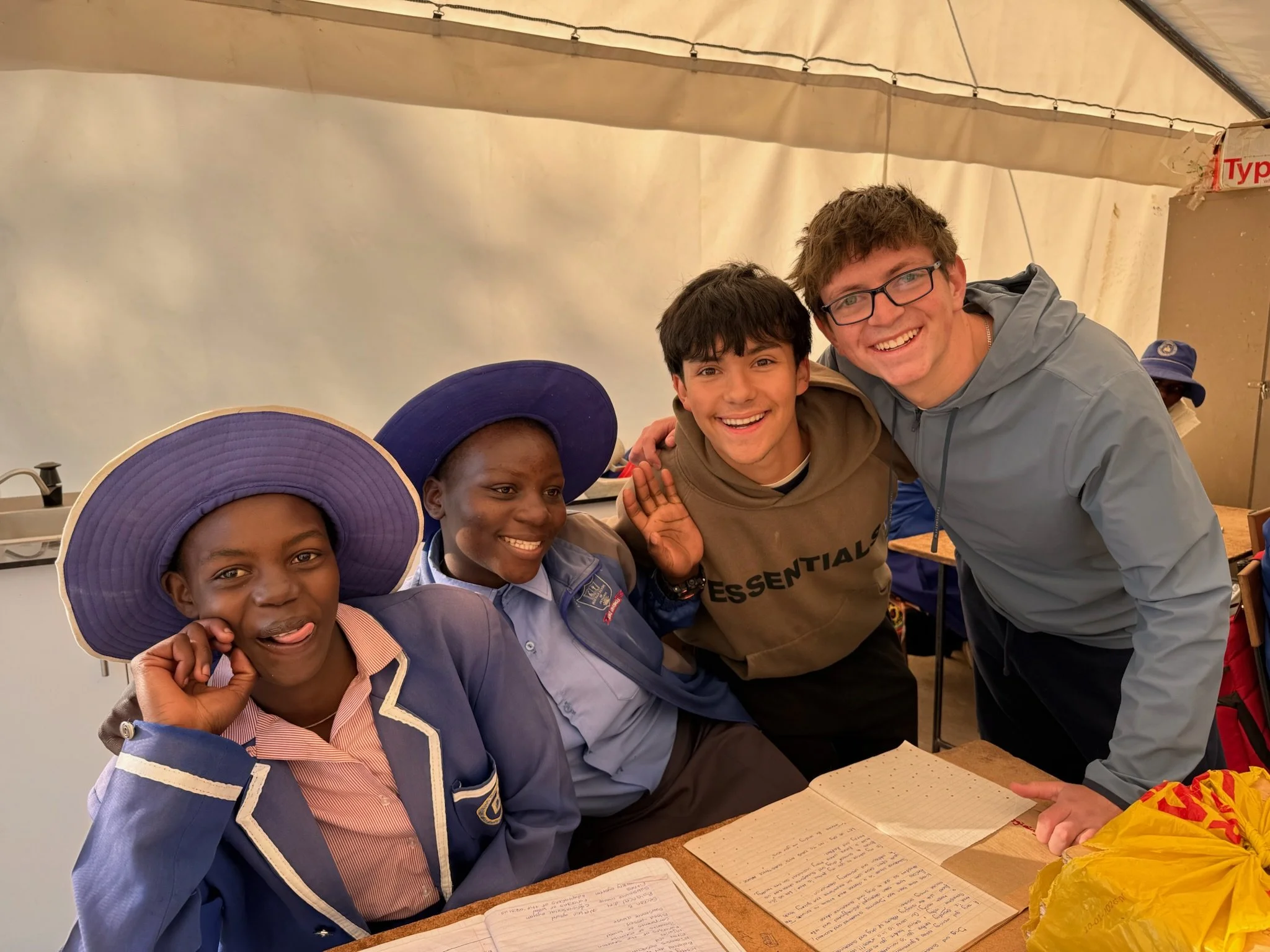 Group of children and a young man smiling and posing for a photo inside a tent or indoor space, with papers on the table in front of them.