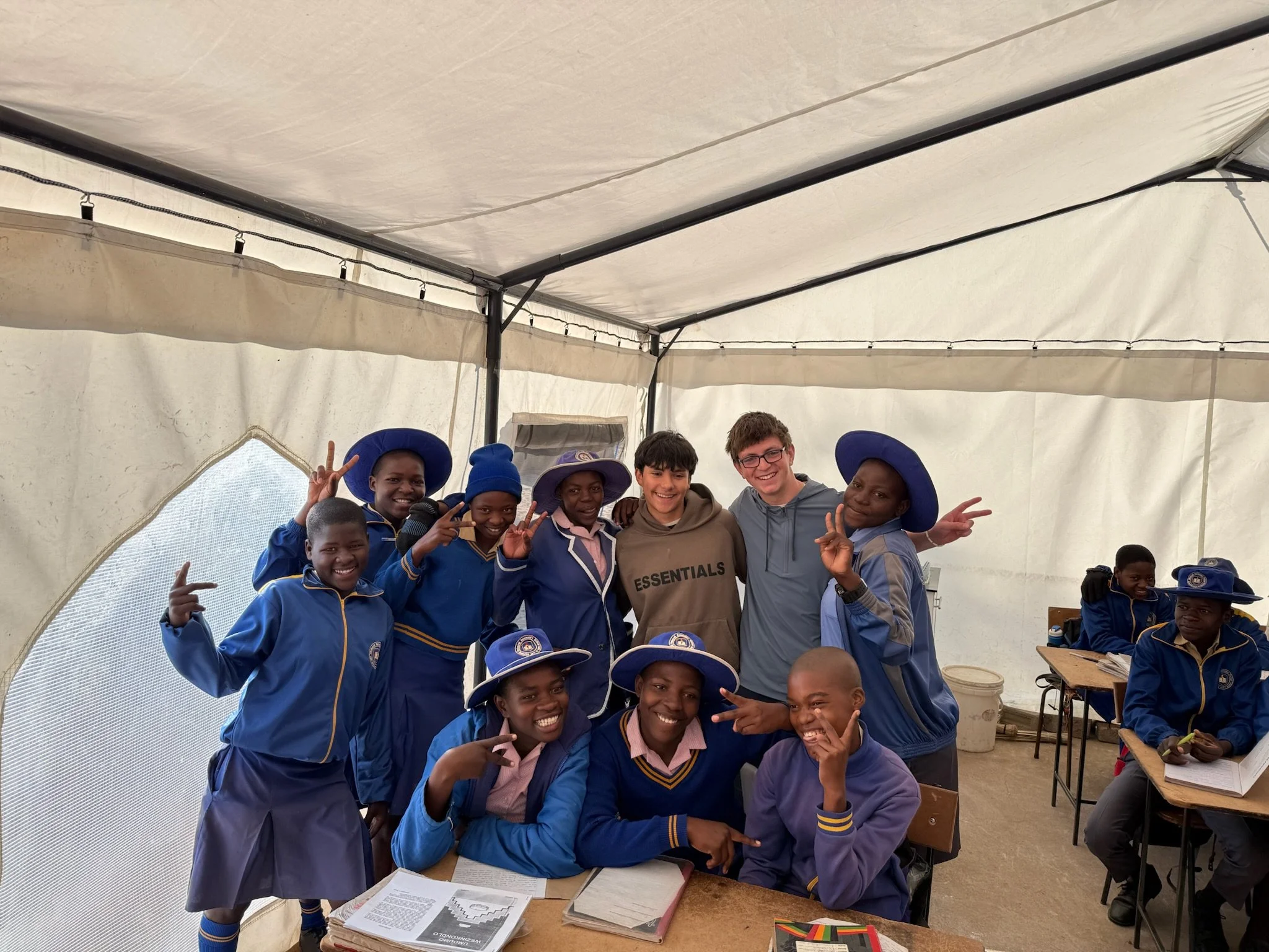 Group of children and teenagers inside a tent, smiling and making peace signs. They are wearing school uniforms and hats. The tent has string lights and tables with books and papers.