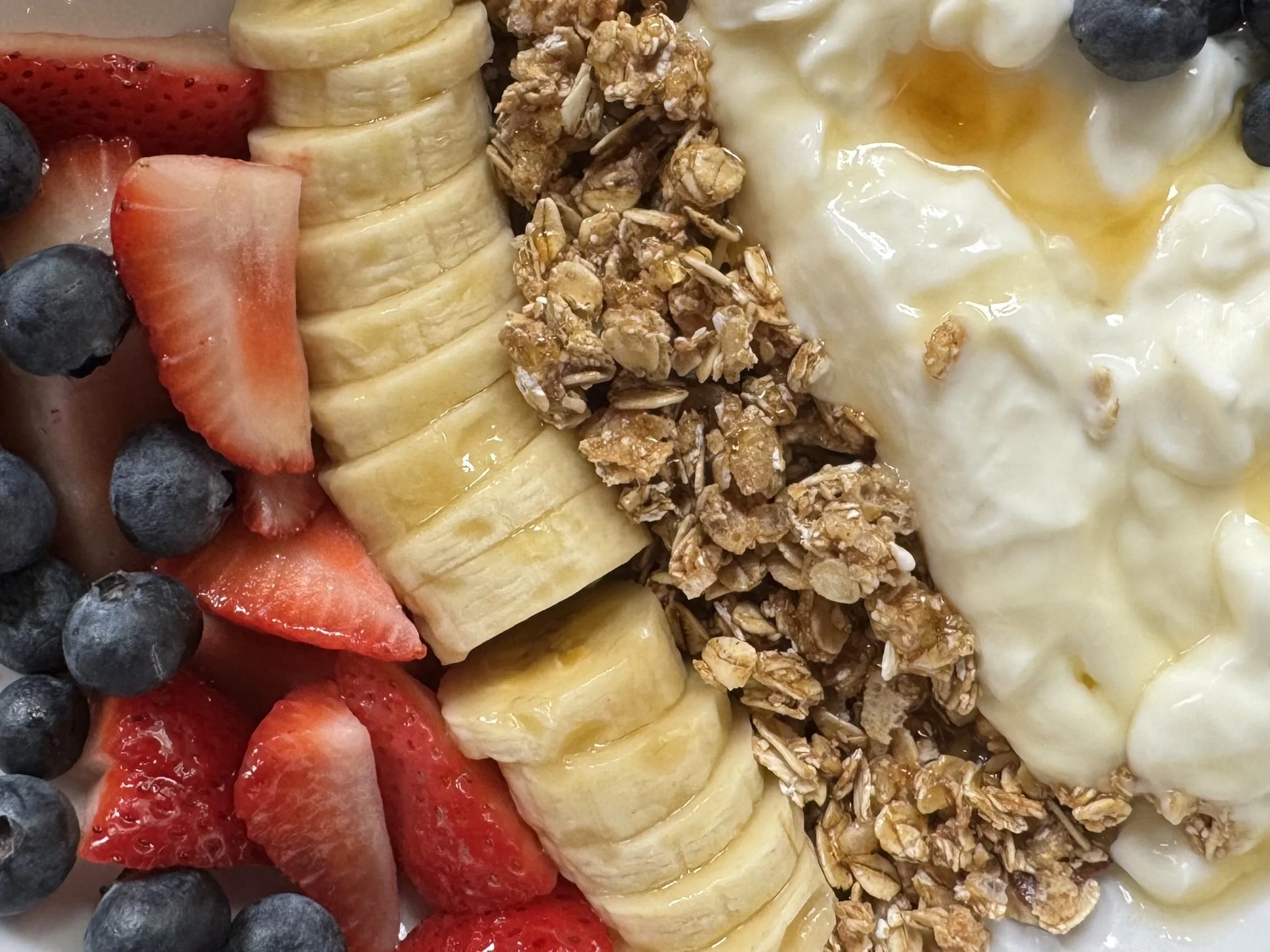Close-up of a fruit and yogurt breakfast bowl featuring blueberries, sliced strawberries, banana slices, granola, and a dollop of yogurt topped with honey.