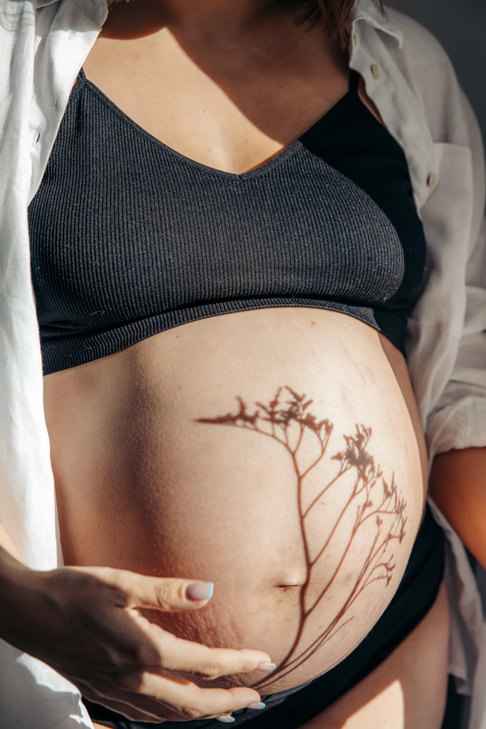 Close-up of a pregnant woman with a delicate shadow on her belly, wearing a black ribbed crop top and an unbuttoned white shirt, with her hands gently cradling her belly.