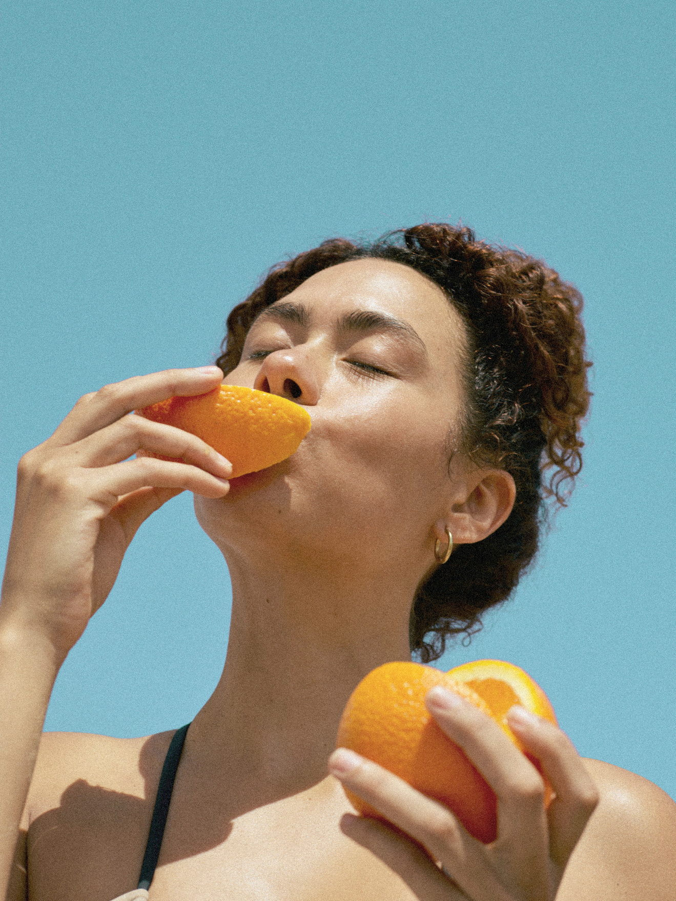 A woman with tied-back curly hair and hoop earrings holding an orange near her face and another orange in her other hand, against a clear blue sky.