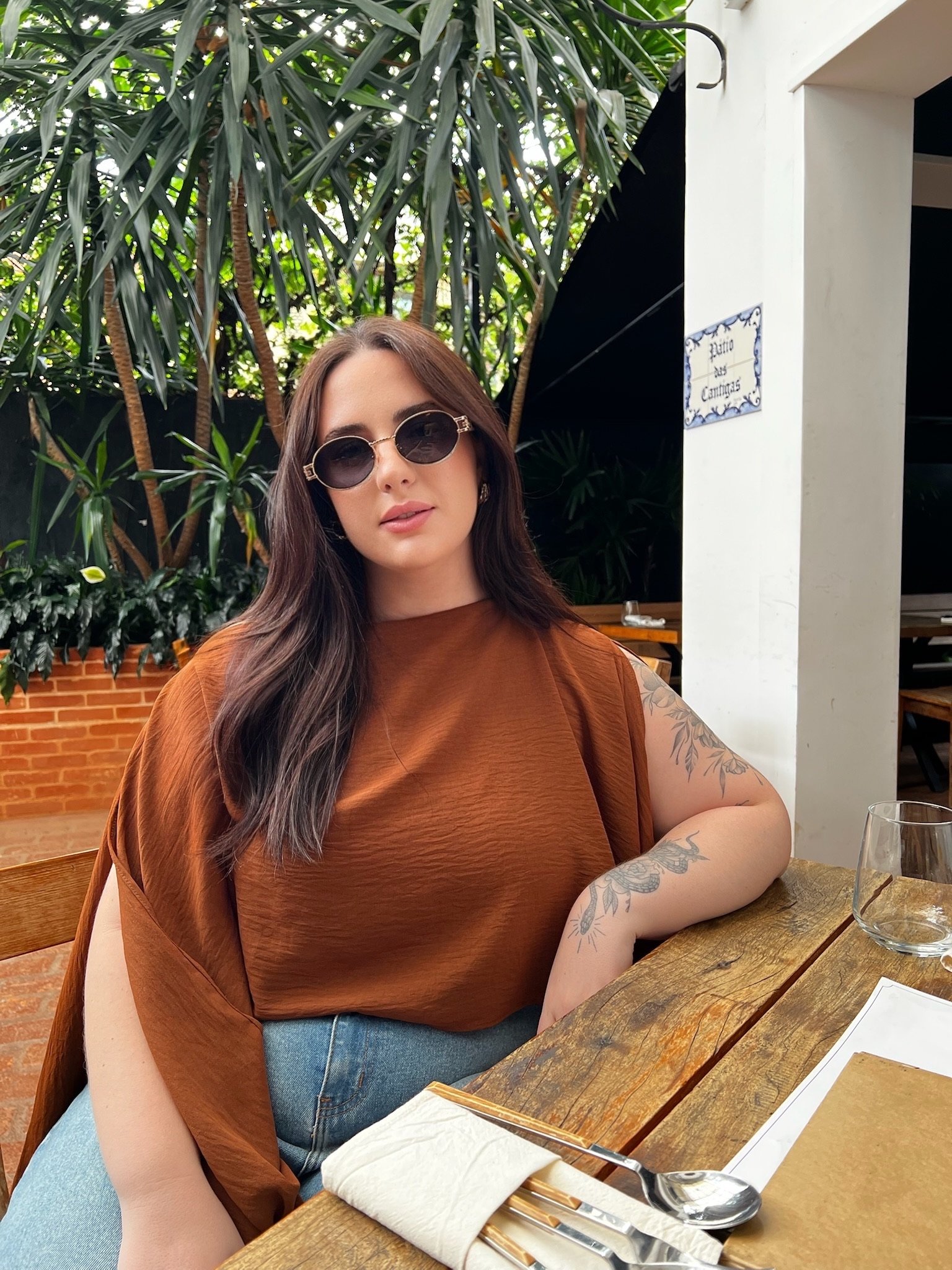 A woman with long dark hair, wearing sunglasses and a brown top, sitting at a wooden table in an outdoor setting with lush green plants in the background.