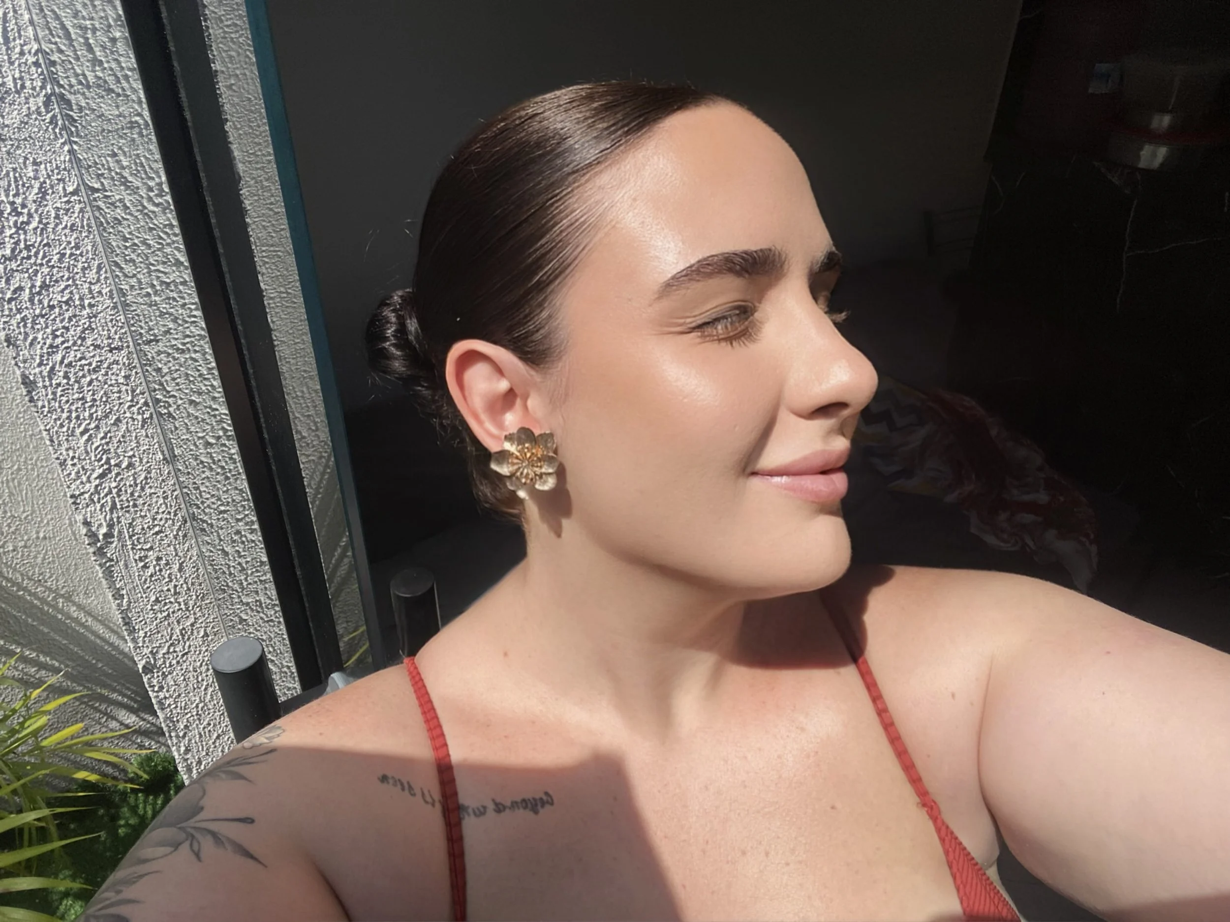 Close-up of a woman's face and shoulder, with a bun hairstyle, wearing gold flower-shaped earrings, a red sleeveless top, and showing a tattoo on her arm, with sunlight on her face.