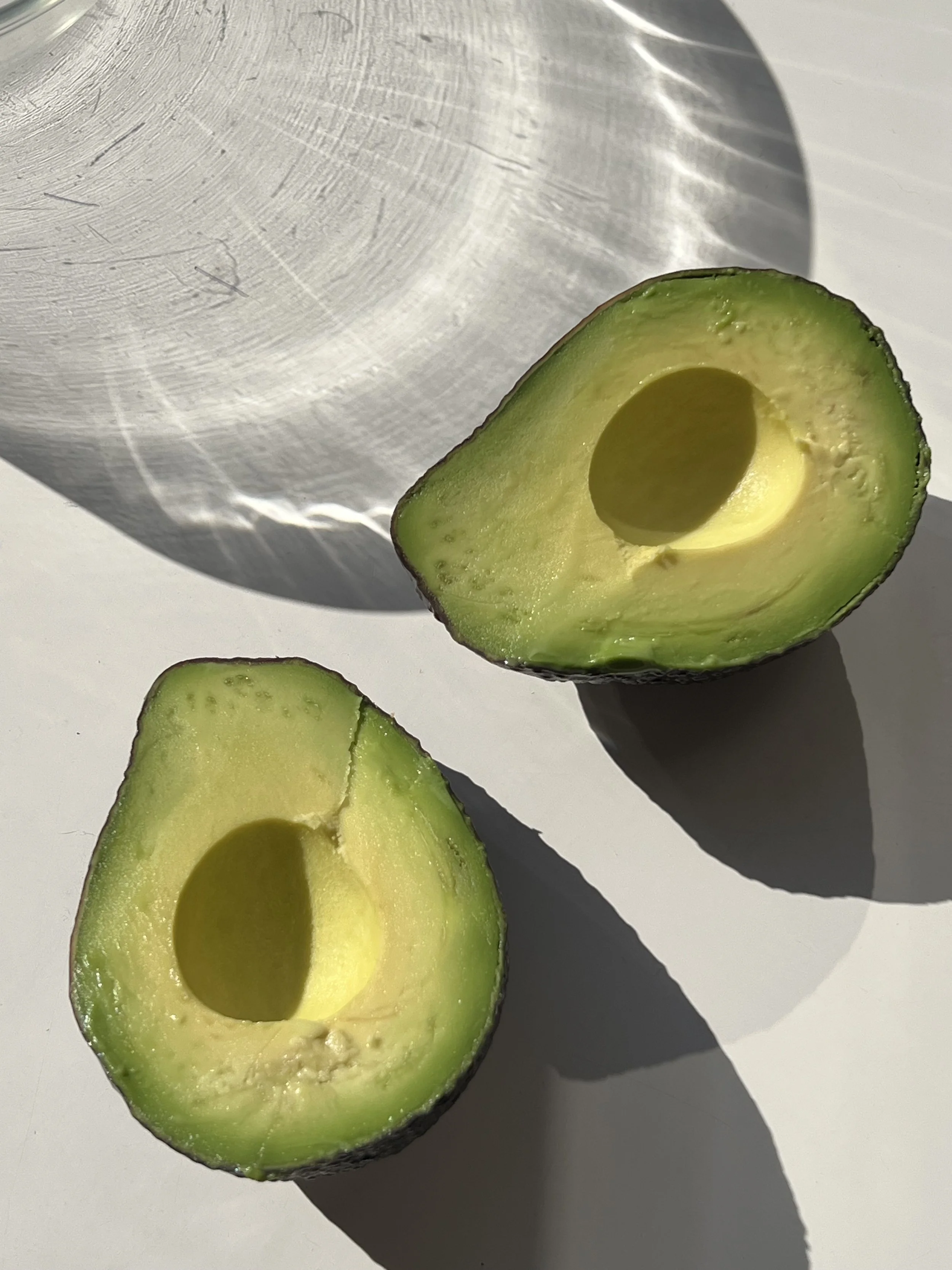 Close-up of a halved avocado with the seed removed, placed on a white surface with a shadow cast by an object in the background.