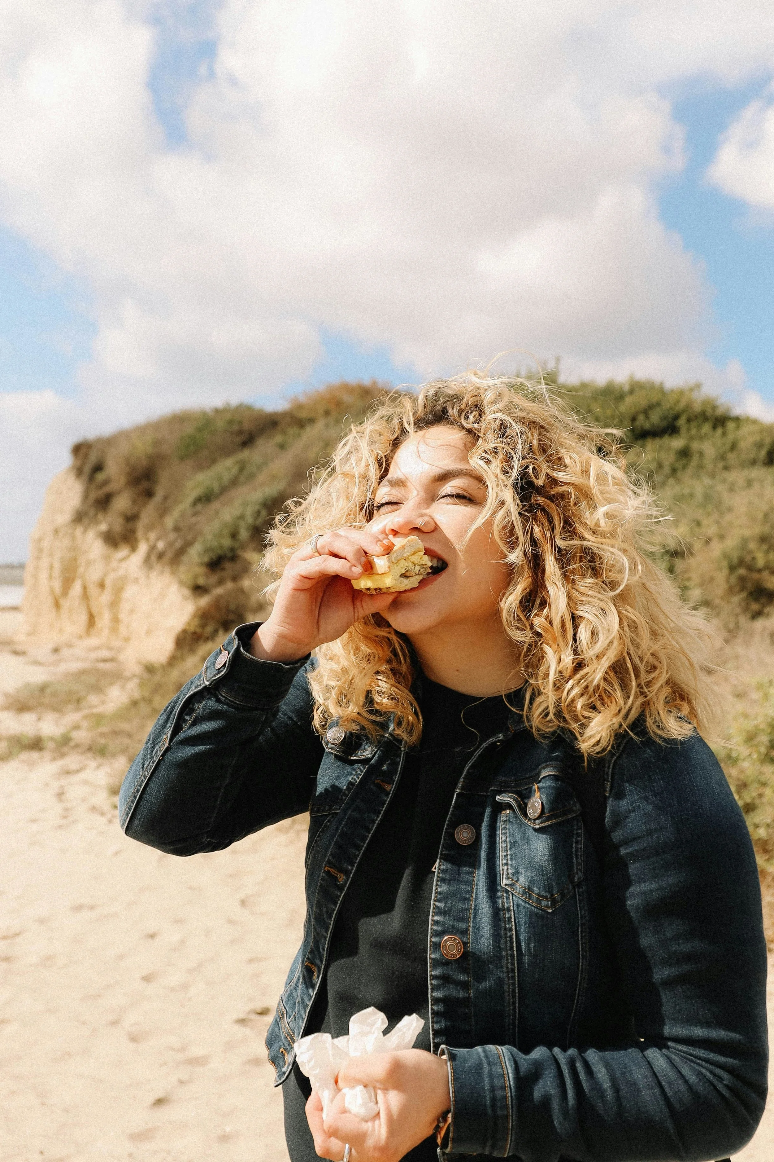 A woman with curly blonde hair enjoying a sandwich outdoors at the beach, wearing a black shirt and denim jacket, with sandy cliffs and a cloudy sky in the background.