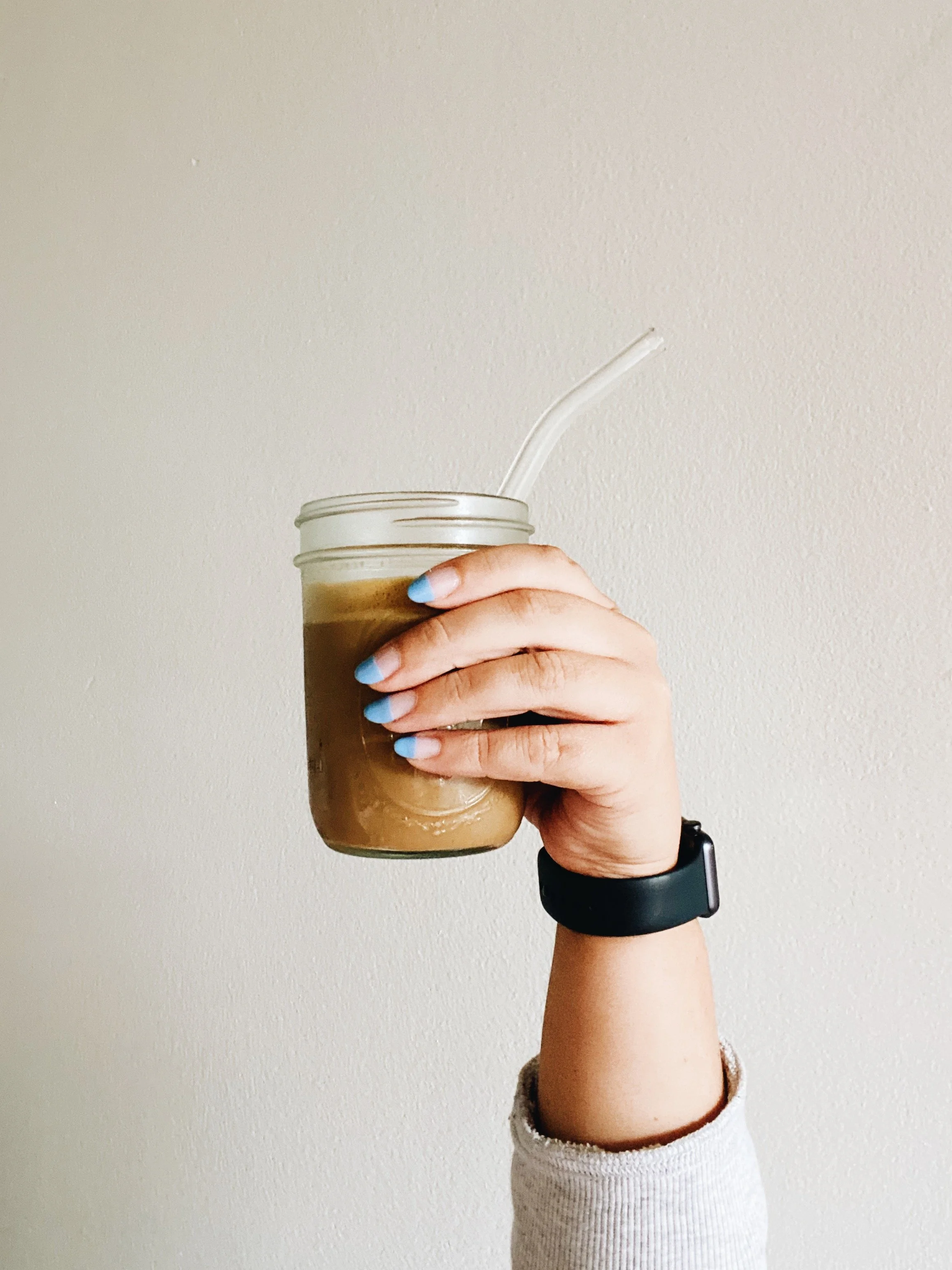 A person holding a glass jar with iced coffee and a white straw, with their fingernails painted light blue, against a plain off-white wall.