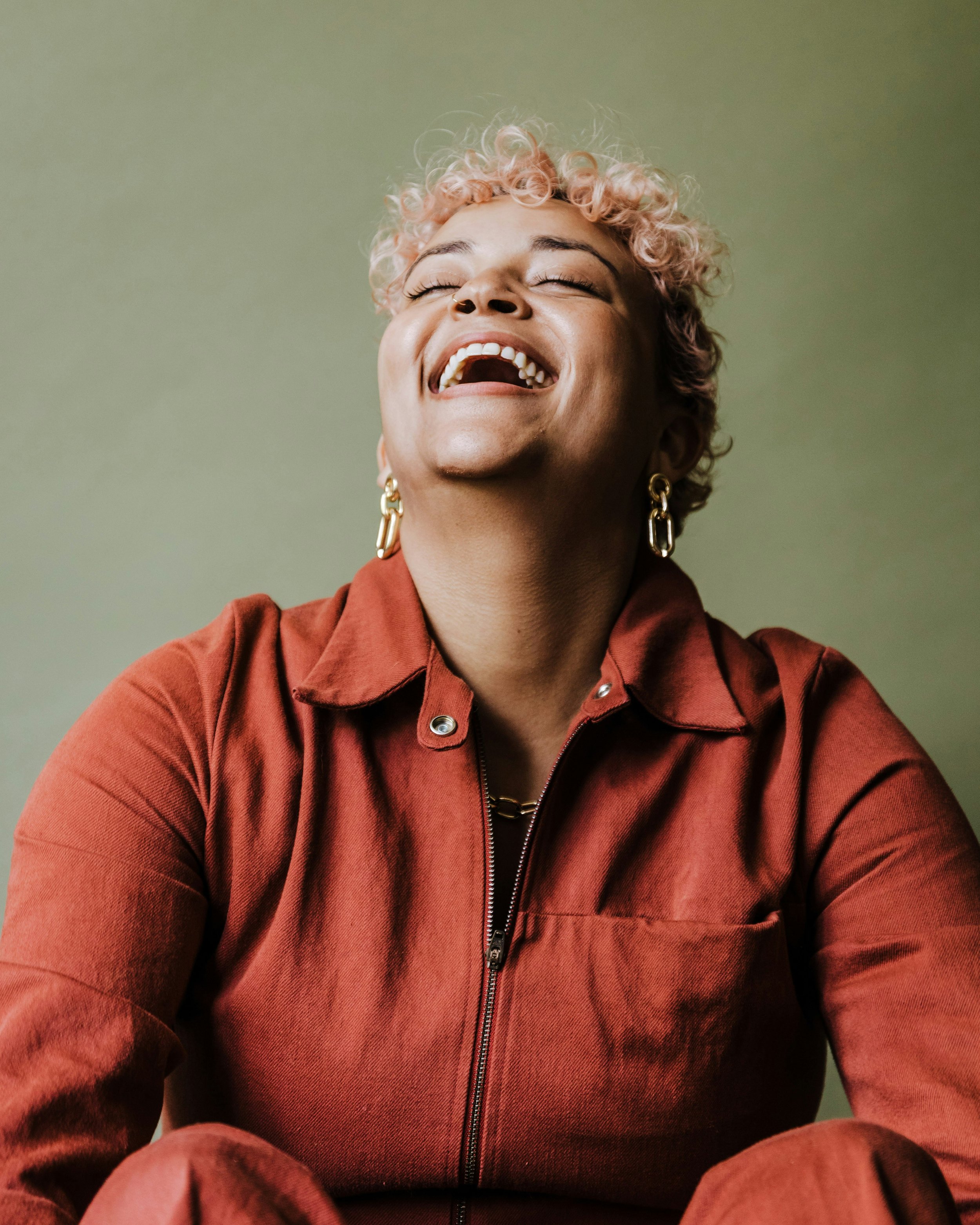 Close-up of a woman with short, curly, light pink hair, wearing a rust-colored jacket and gold earrings, smiling and laughing with her eyes closed against a plain, muted green background.