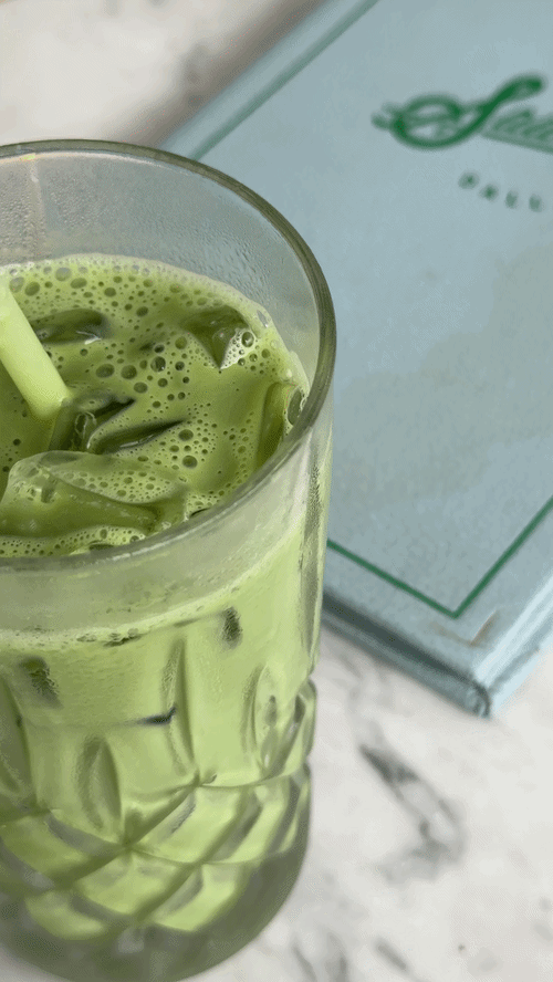 A tall glass of green matcha smoothie with ice cubes and a green straw on a marble surface, with a box of matcha tea in the background.