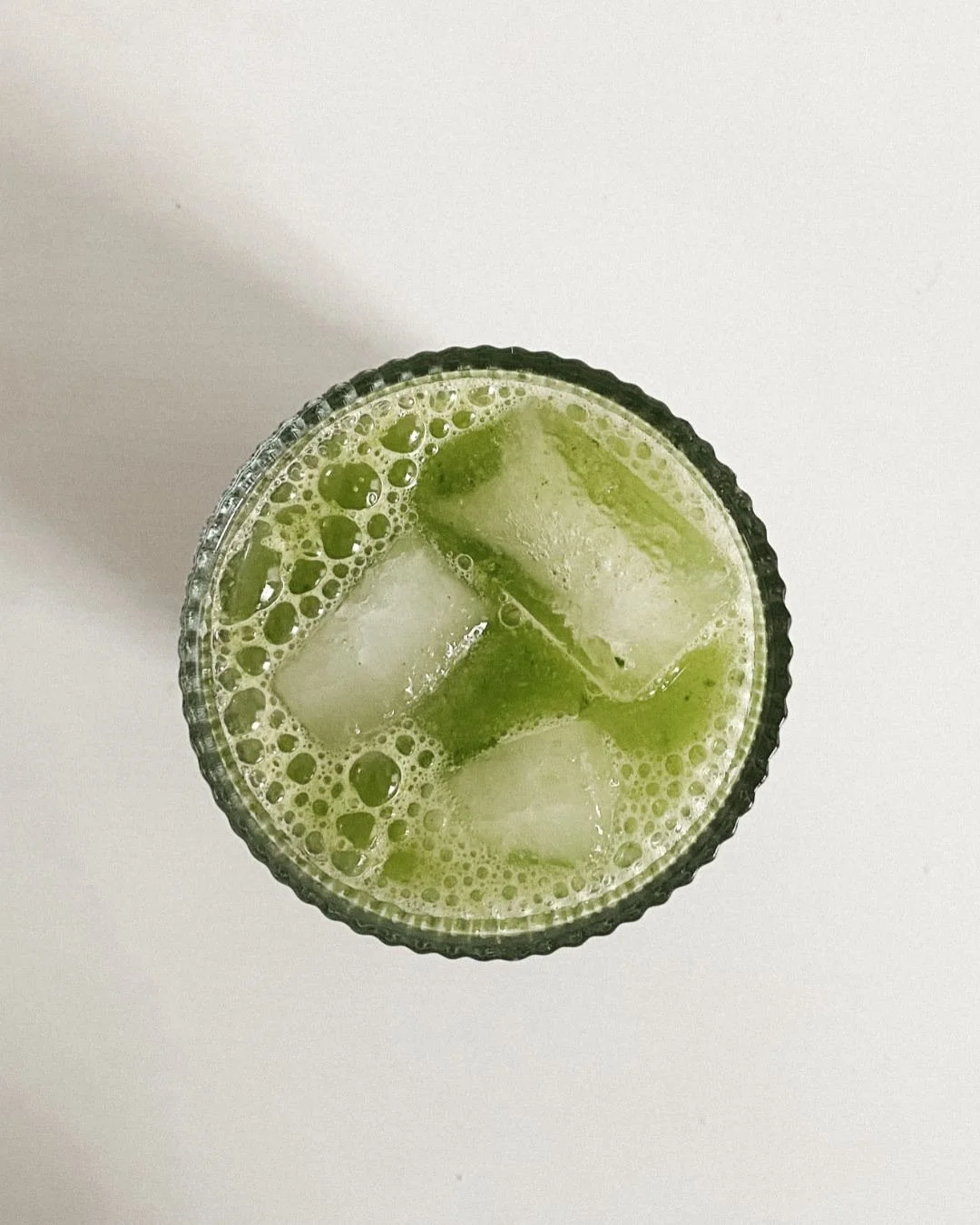 Top-down view of a glass of green beverage with ice cubes on a white surface.