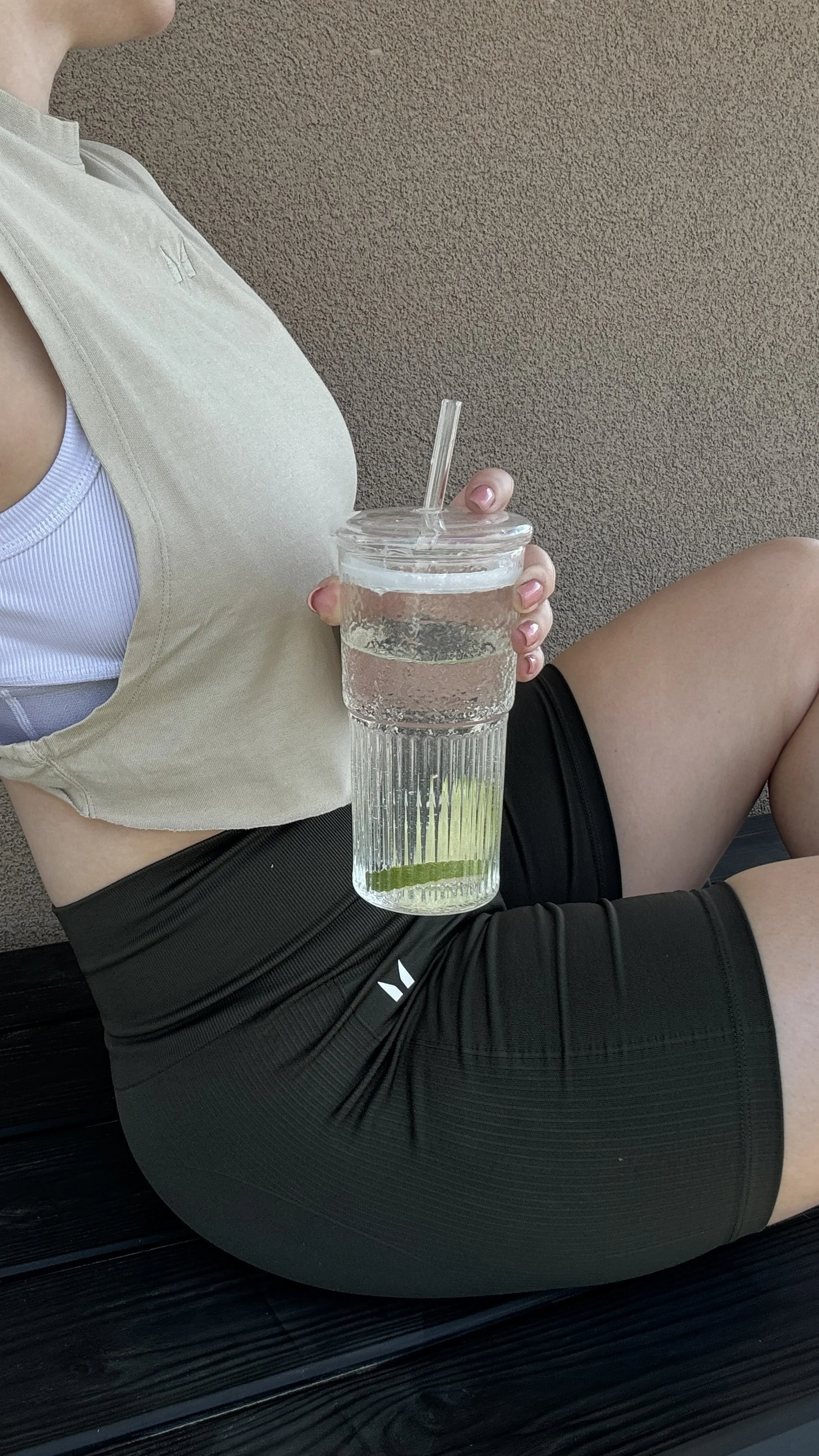 A woman in athletic wear sitting outside against a textured brown wall, holding a large mason jar with a lid and straw containing water and lime slices.