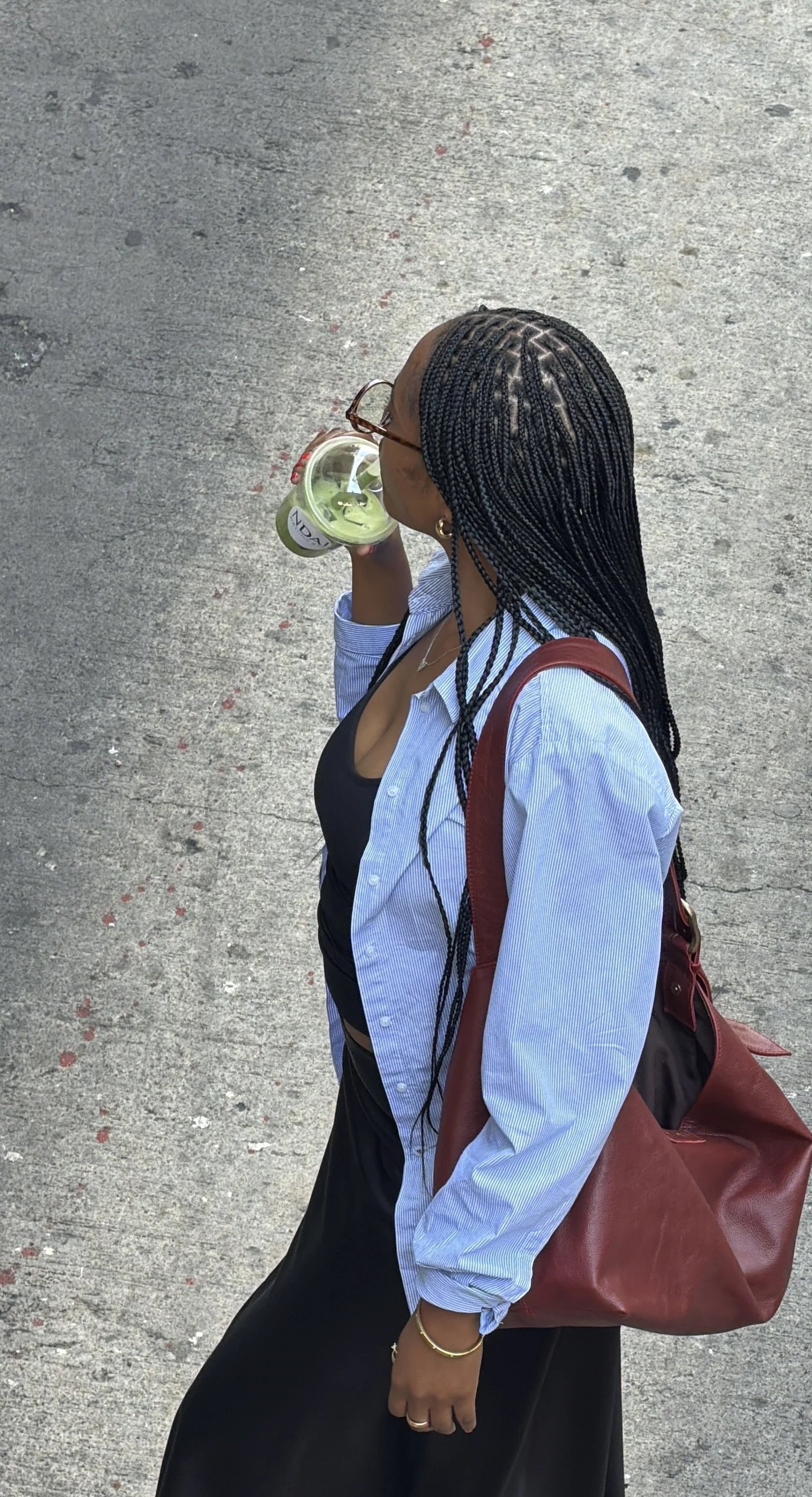 A woman with braided hair, wearing glasses, a black top, a blue and white striped shirt, and a black skirt, is walking on a concrete pathway while drinking a green beverage from a plastic cup with a straw. She is carrying a large brown leather bag on her shoulder and has a gold bracelet and rings on her right hand.