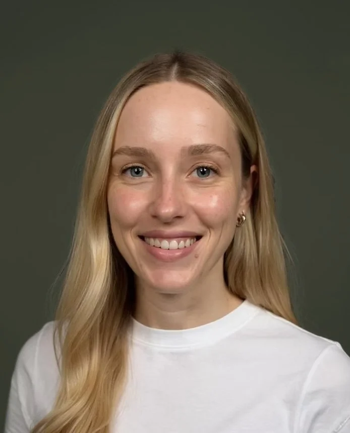 A young woman with long blonde hair, blue eyes, and a warm smile, wearing a white shirt and earrings, against a plain dark background.