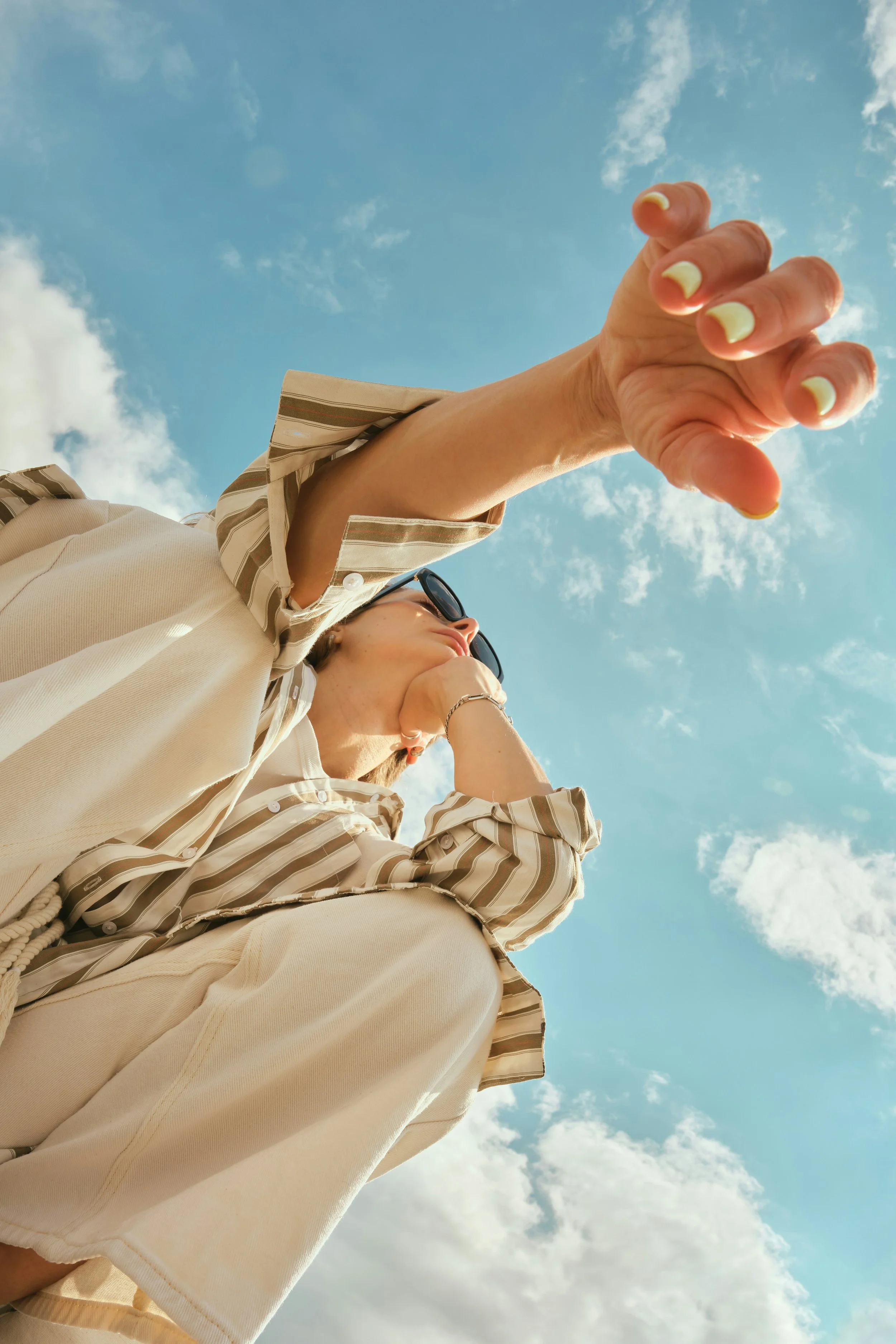 A woman leans over the camera, wearing a beige outfit with stripes. A blue sky with white clouds is behind her.