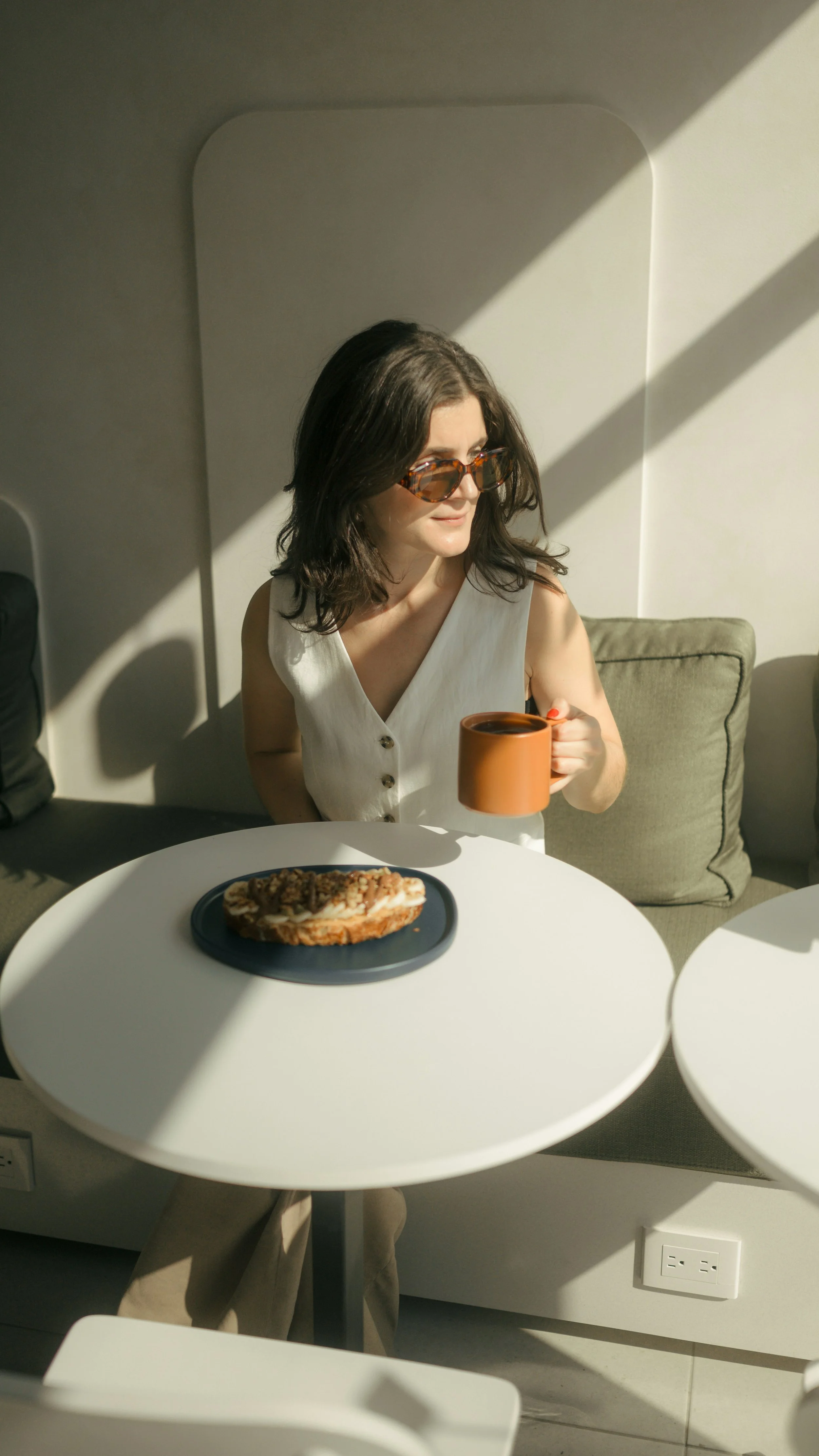 A woman with dark hair and sunglasses sitting at a white table, holding a cup of coffee or tea, with a plate of food in front of her, in a bright room with sunlight casting shadows.