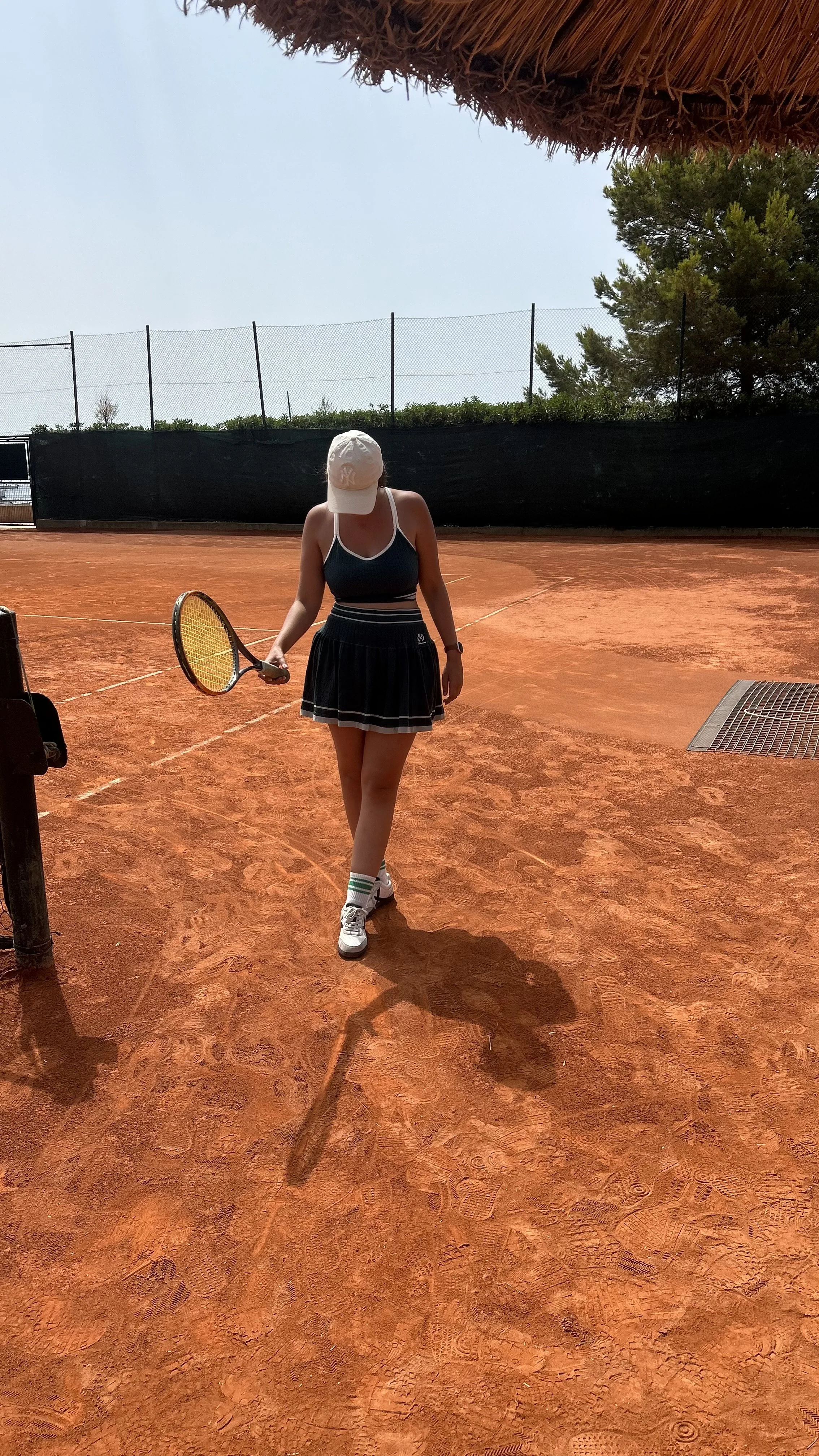 A woman in tennis attire, holding a tennis racket, walking on a clay tennis court with feet partially visible, under a canopy with trees and a black windscreen in the background.