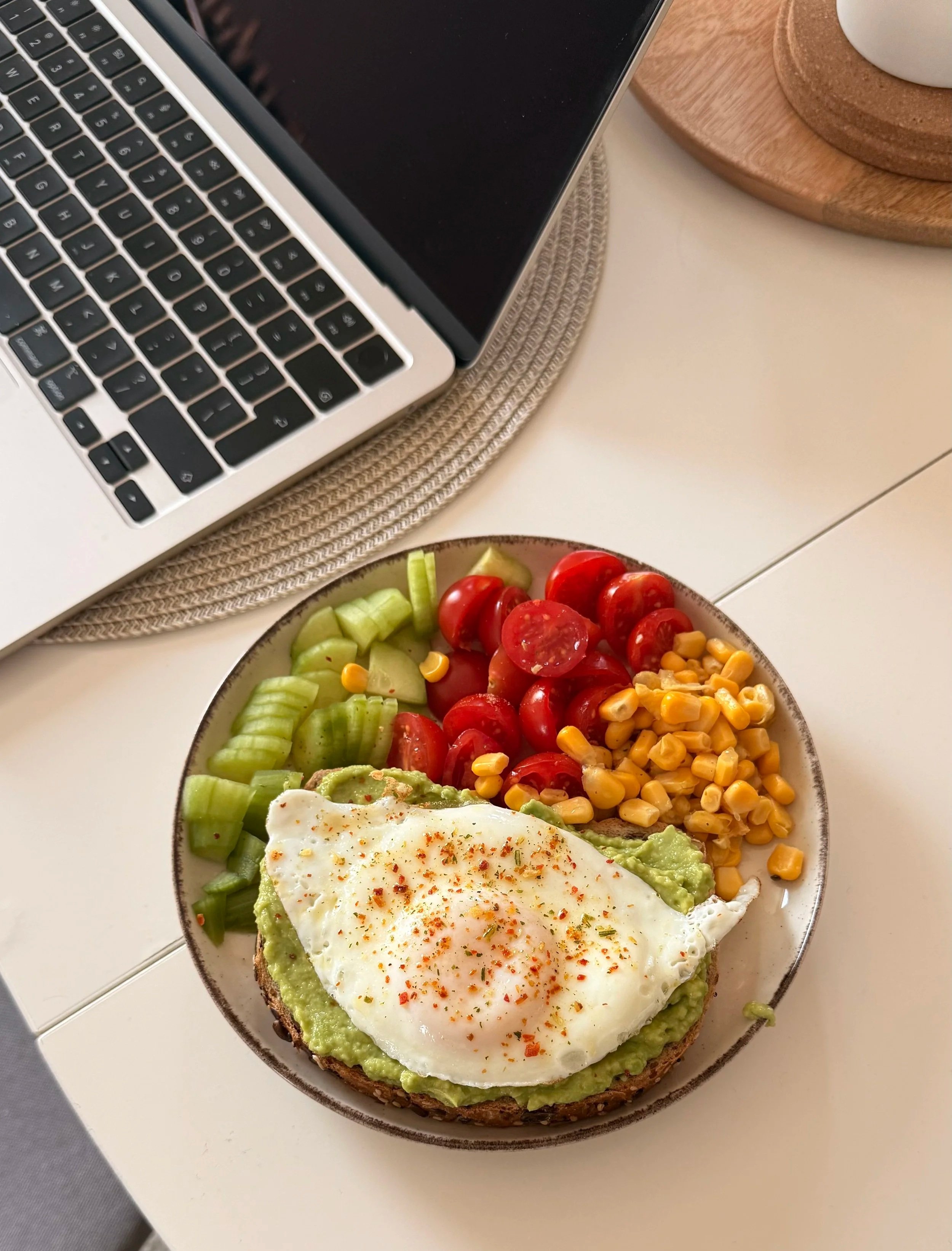 A plate of food with sliced cherry tomatoes, corn, sliced cucumber, and avocado on toasted bread topped with a fried egg and sprinkled red pepper