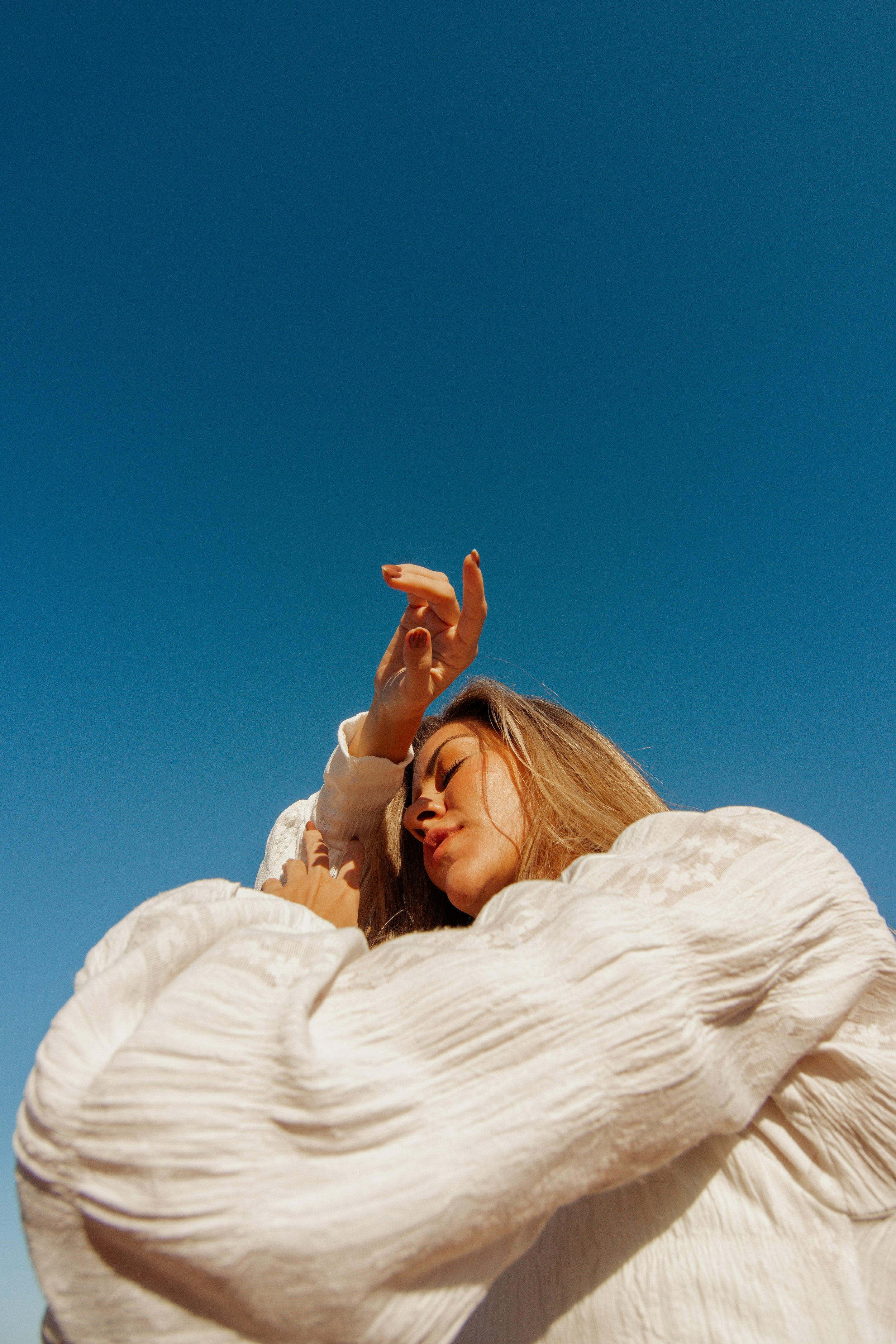 A woman with blonde hair is resting with her eyes closed, wearing a white, long-sleeved, textured dress. She is holding her right arm with her left hand. The background shows a clear blue sky, and part of her face and hand are visible.