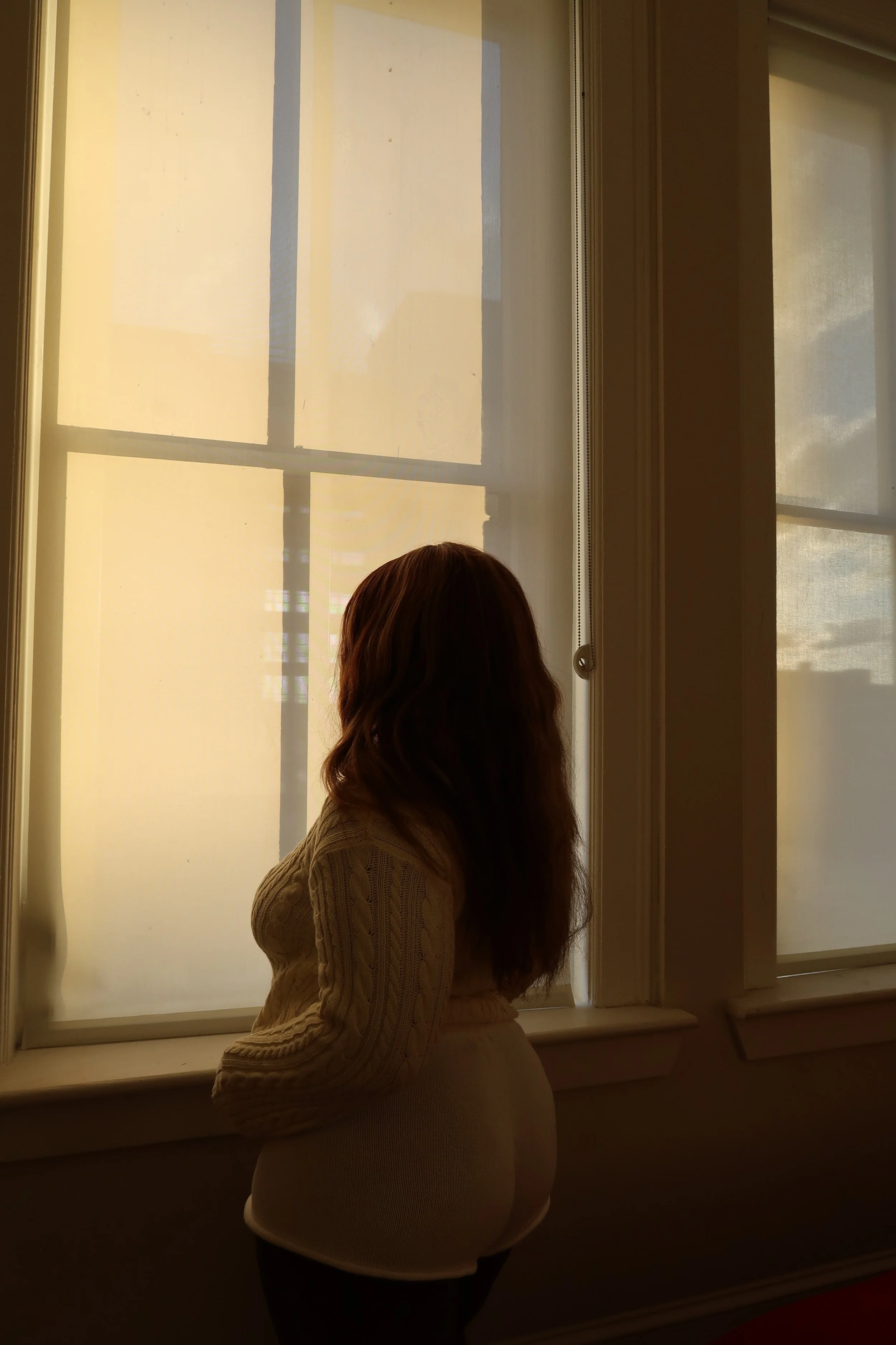 A woman standing in front of a window with yellow blinds, looking outside. She is wearing a beige sweater and has long wavy hair.