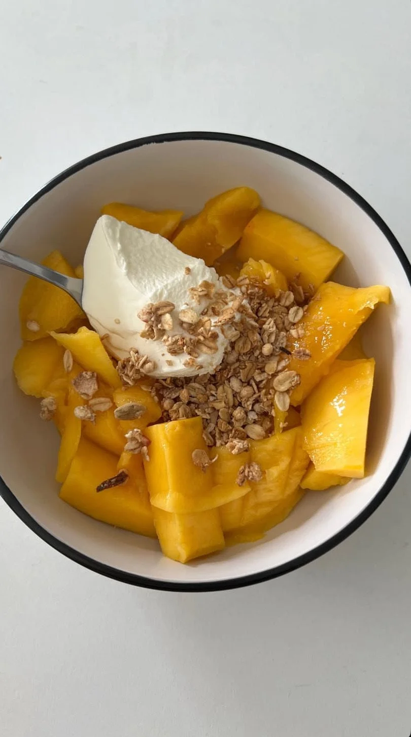 Bowl of chopped yellow mango, topped with a dollop of yogurt, granola, and a spoon.