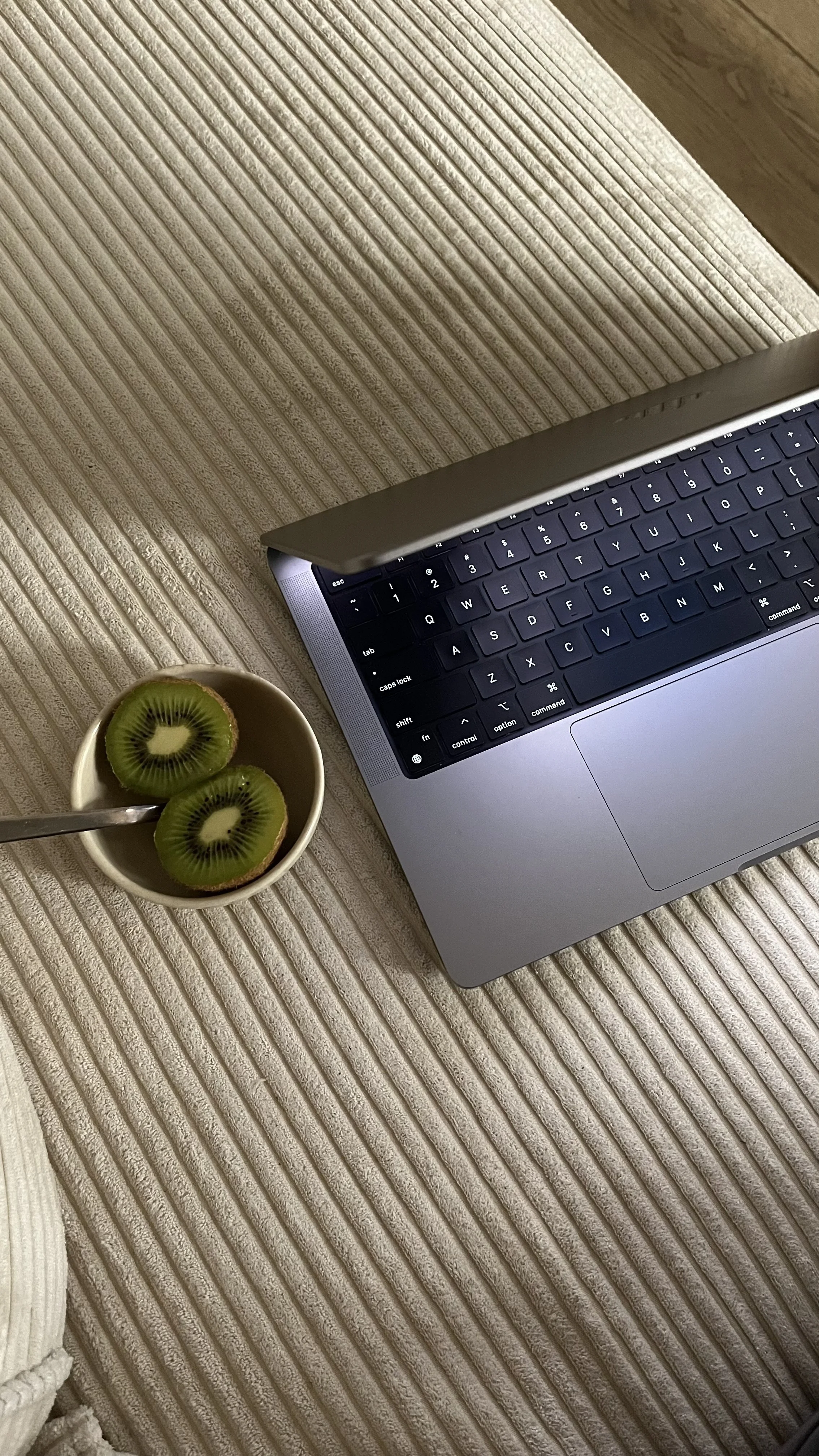 Laptop on a beige textured surface next to a cup with two slices of kiwi.