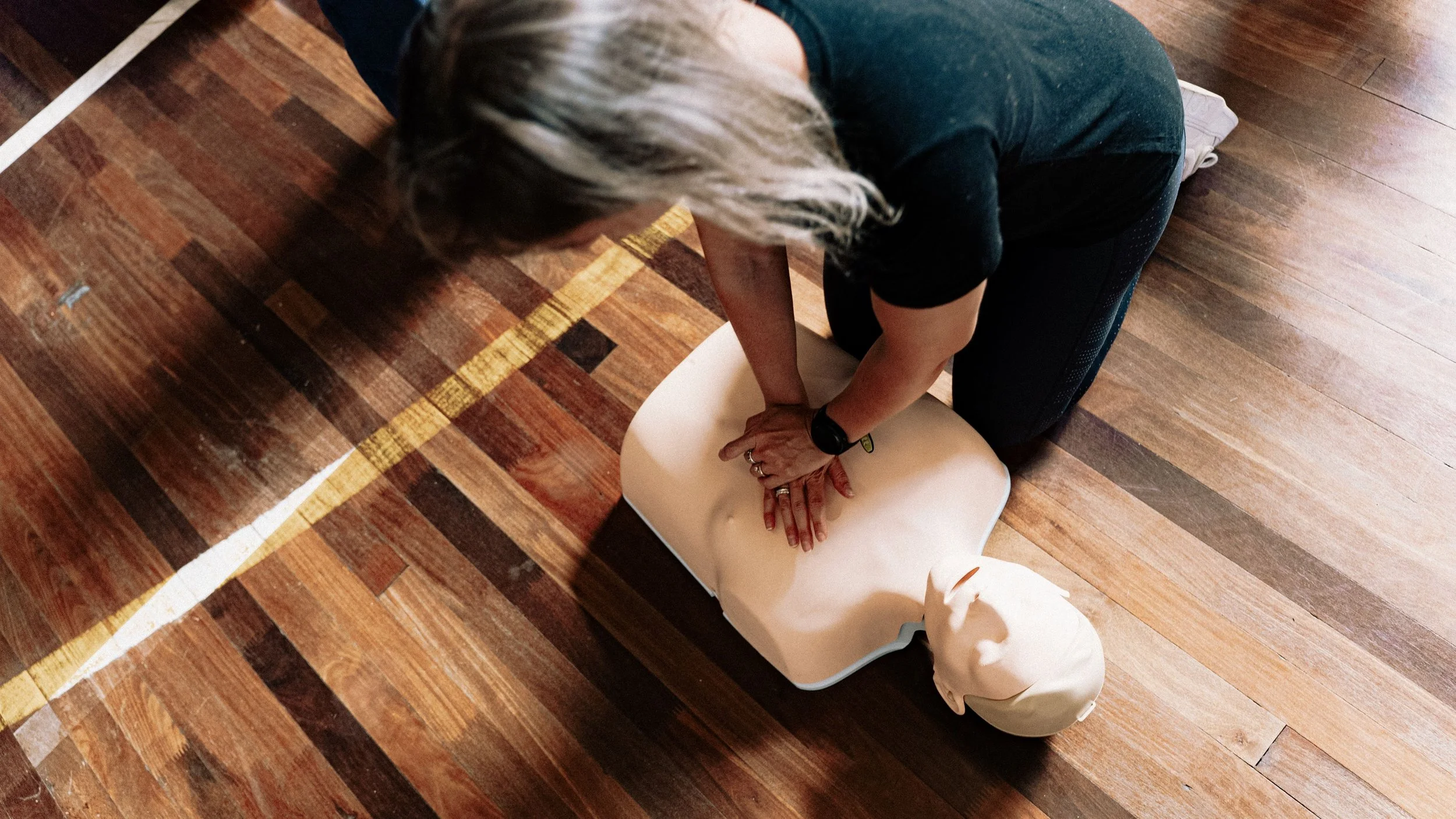 Person practicing CPR on a training mannequin in a room with wooden floors.