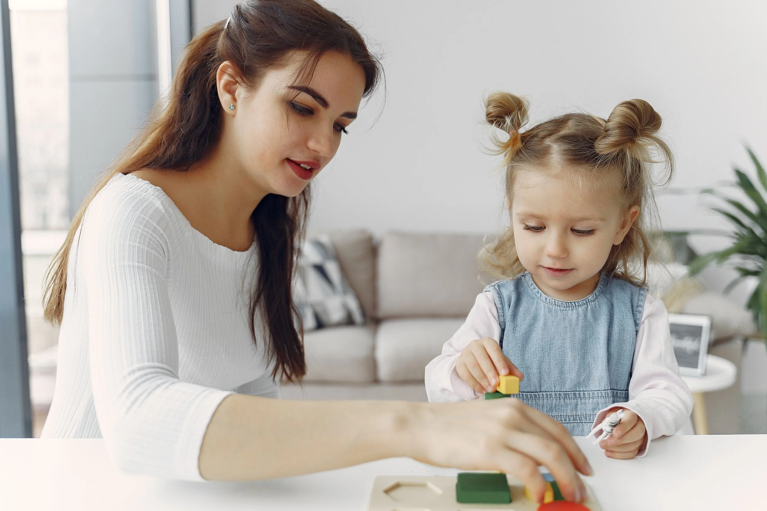 A young woman and a young girl playing with colorful building blocks at a white table in a bright, modern living room.