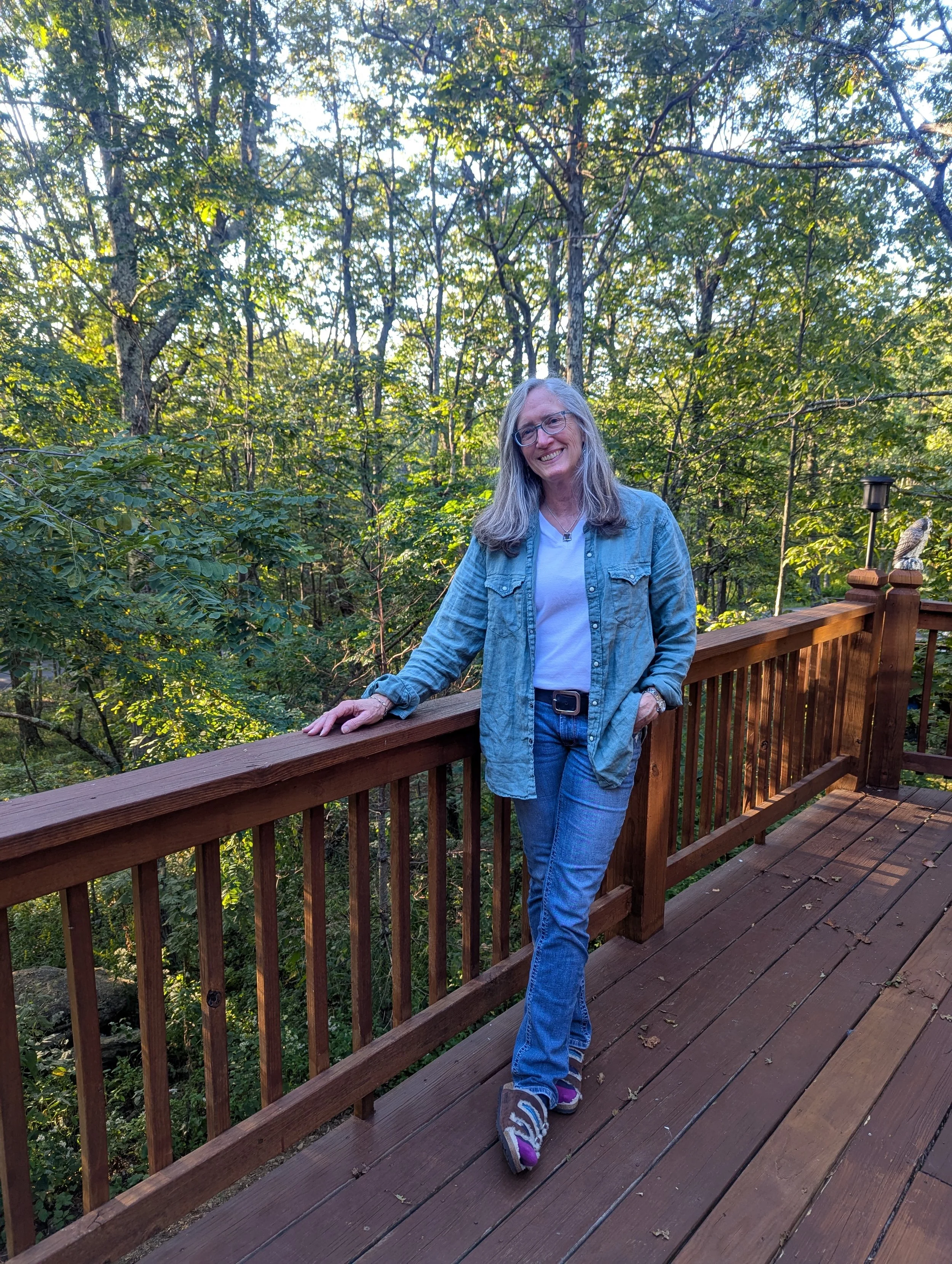 A smiling woman with long gray hair and glasses posing on a wooden deck in a lush green forest, wearing a denim jacket and white shirt.