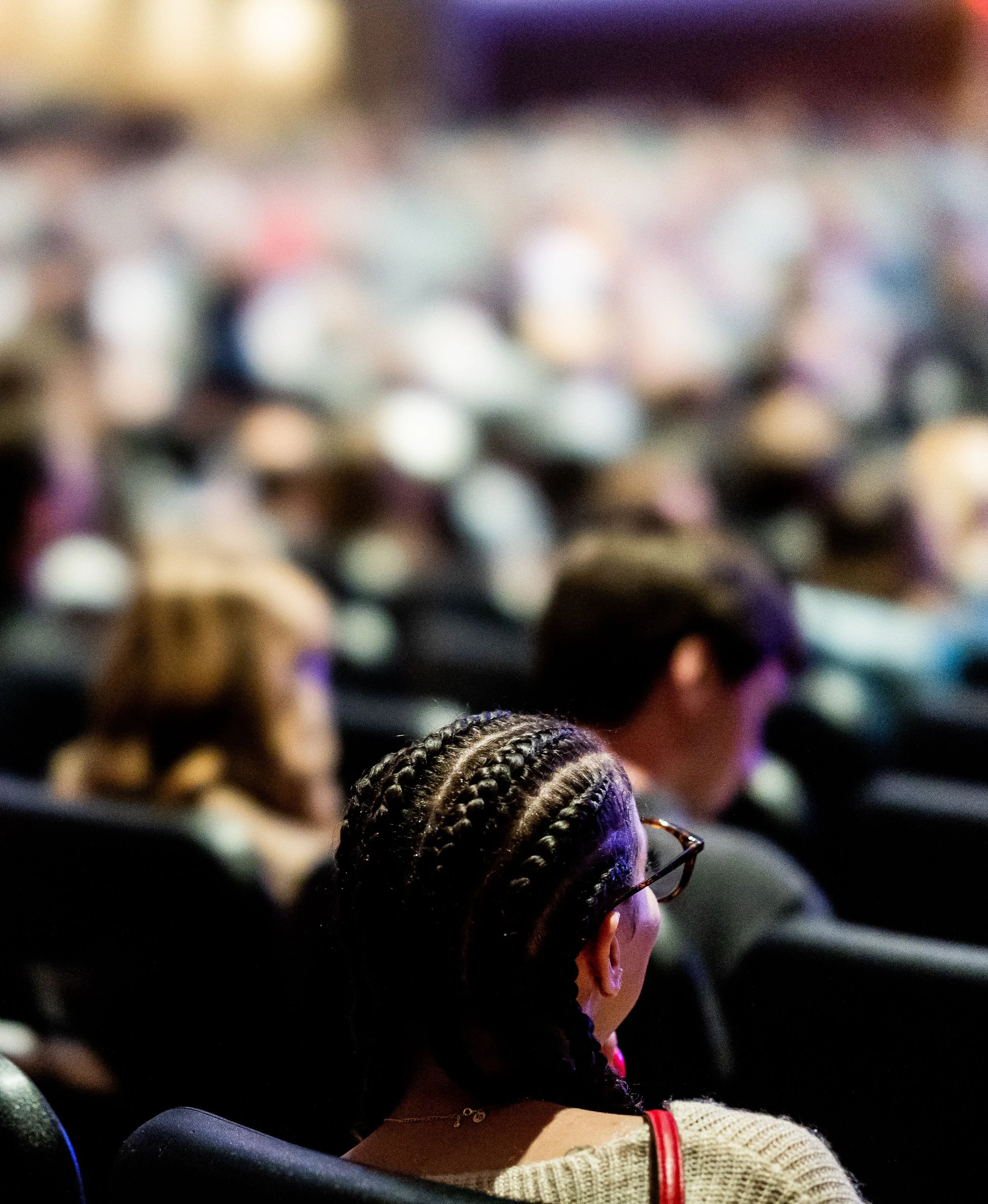 A woman with dark braided hair wearing glasses and a white top with red accents, sitting in an auditorium with dark seats, attending an event or performance with a blurred background of other attendees.