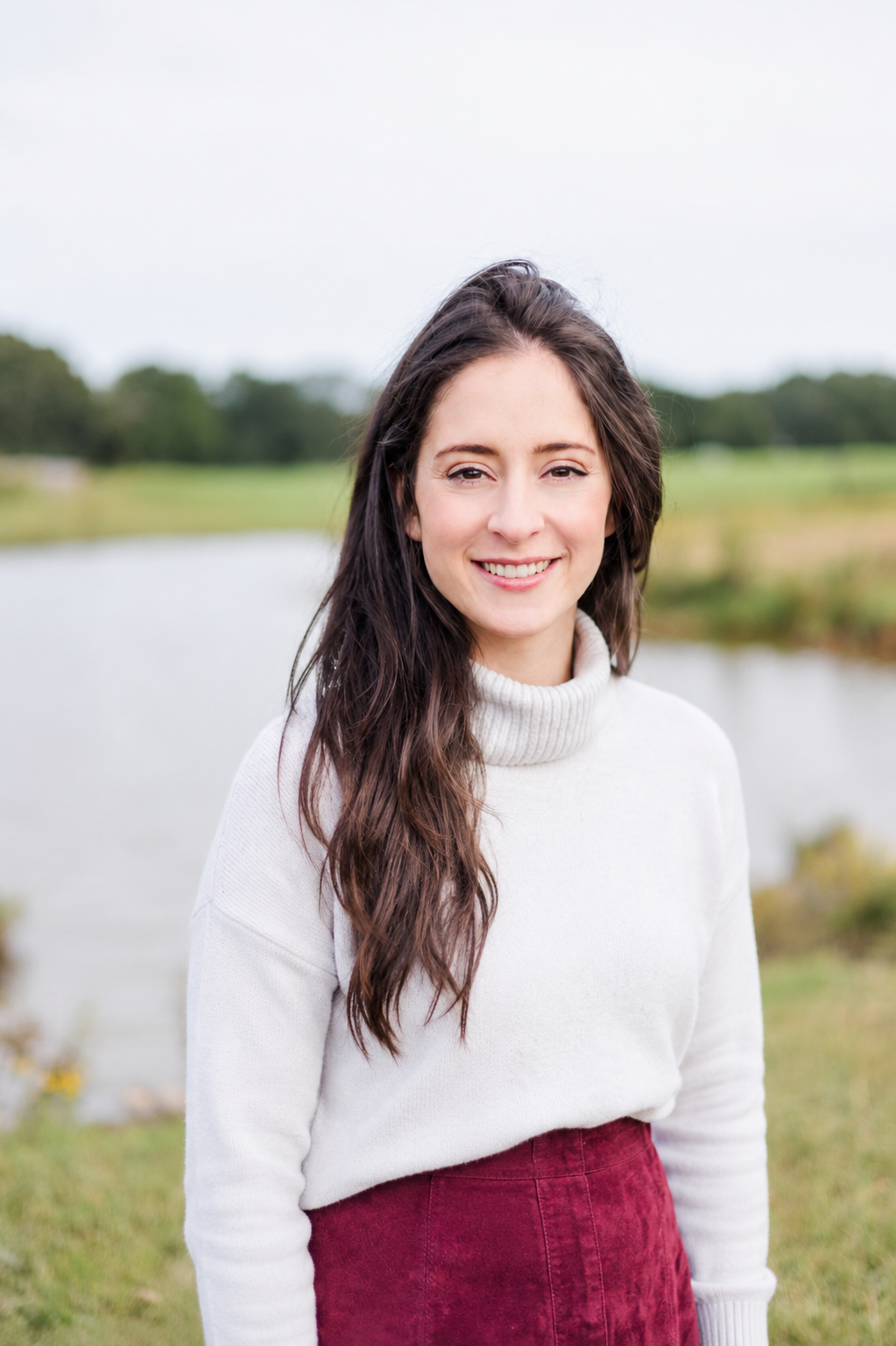 A young woman with long dark hair smiling outdoors near a body of water, wearing a cream turtleneck sweater and a red skirt.