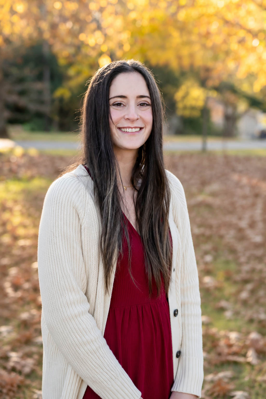 A woman with long dark hair wearing a red dress and beige cardigan smiling outdoors during autumn.