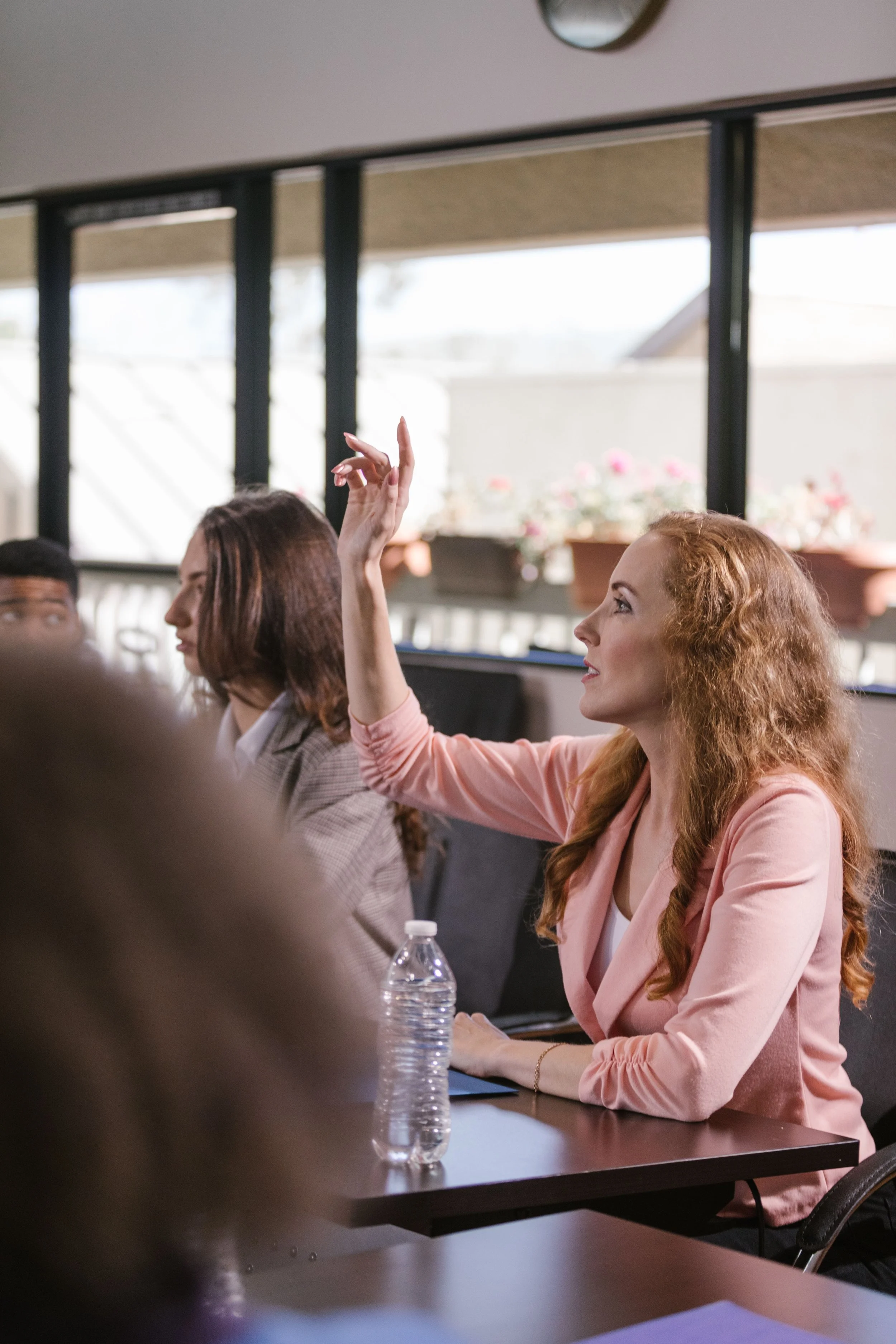 A woman with long curly red hair wearing a pink blazer and white top, sitting at a conference table in a meeting room, raising her hand. Other people are seated around the table, including a woman with straight brown hair and a man with short black hair. There is a bottle of water on the table and a laptop partially visible. The background shows a window with potted flowers and sunlight coming in.