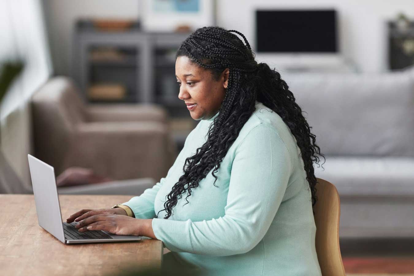 Woman with braided hair sitting at a wooden table using a laptop in a living room.